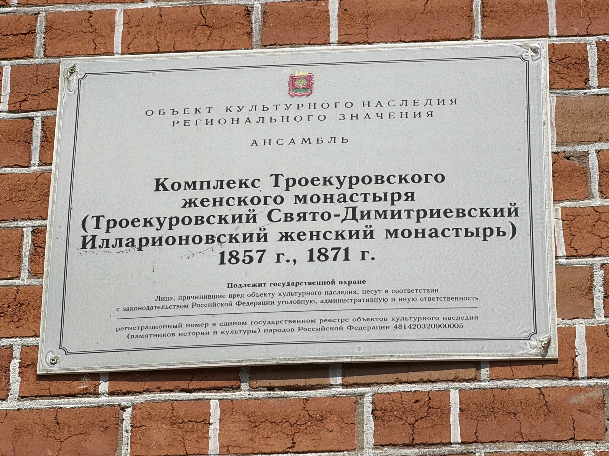 The image shows a plaque mounted on a brick wall. The plaque is rectangular with a light gray background and a decorative border. The text on the plaque is in Russian and appears to be an official designation of a cultural heritage site.
The top of the plaque reads "ОБЪЕКТ КУЛЬТУРНОГО НАСЛЕДИЯ РЕГИОНАЛЬНОГО ЗНАЧЕНИЯ" which translates to "OBJECT OF CULTURAL HERITAGE OF REGIONAL SIGNIFICANCE". Below this is "АНСАМБЛЬ" meaning "ENSEMBLE".
The main body of the text states "Комплекс Троекуровского женского монастыря (Троекуровский Свято-Димитриевский Илларионовский женский монастырь) 1857 г., 1871 г." This translates to "Complex of the Troekurovo Women's Monastery (Troekurovo Svyato-Dimitrievsky Illarionovsky Women's Monastery) 1857, 1871".
Further down, it says "Подлежит государственной охране" which means "Subject to state protection". This is followed by legal information regarding responsibility for damage and a registration number "481420320900005".
The brick wall is made of red bricks with visible mortar lines. The plaque is attached to the wall with visible screws or rivets at the corners. The lighting suggests it is daytime, and the overall scene is static, depicting an informational sign. The location is implied to be in Troekurovo, Russia, due to the name of the monastery.