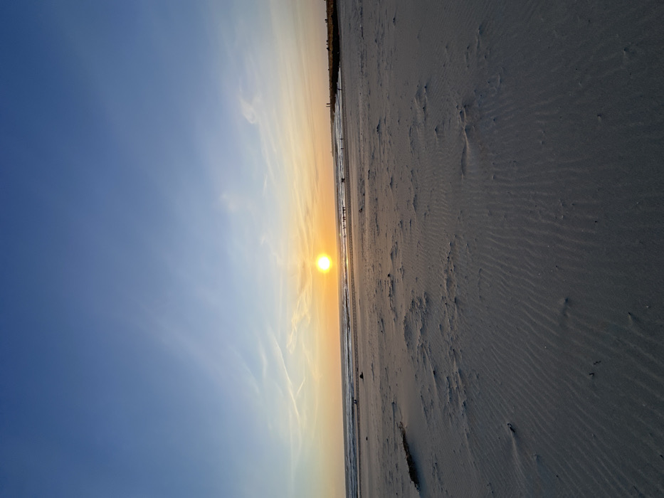 The image displays a coastal scene at sunset in Gafanha da Nazaré, Portugal. The sky occupies the upper two-thirds, showing a gradient from deep blue overhead to a vibrant orange and yellow hue near the horizon. Wispy cirrus clouds and faint contrails are visible in the upper and mid-sky. A prominent, bright, orange-yellow sun is positioned slightly above the horizon, directly over the ocean. The ocean surface exhibits dark blue-grey waves, reflecting the sun's light in the distance, breaking gently along the shoreline. The lower half of the image features a wide expanse of light grey-brown sand, marked with distinct, wind-formed ripple patterns and irregular mounds. A dark, elongated object, consistent with driftwood or seaweed, is partially visible in the mid-left foreground. Multiple small, dark human silhouettes are observable along the waterline and further down the beach towards the right. A larger cluster of individuals is discernible near a distant dark structure or landmass on the far right horizon. Individuals appear to be walking or standing.