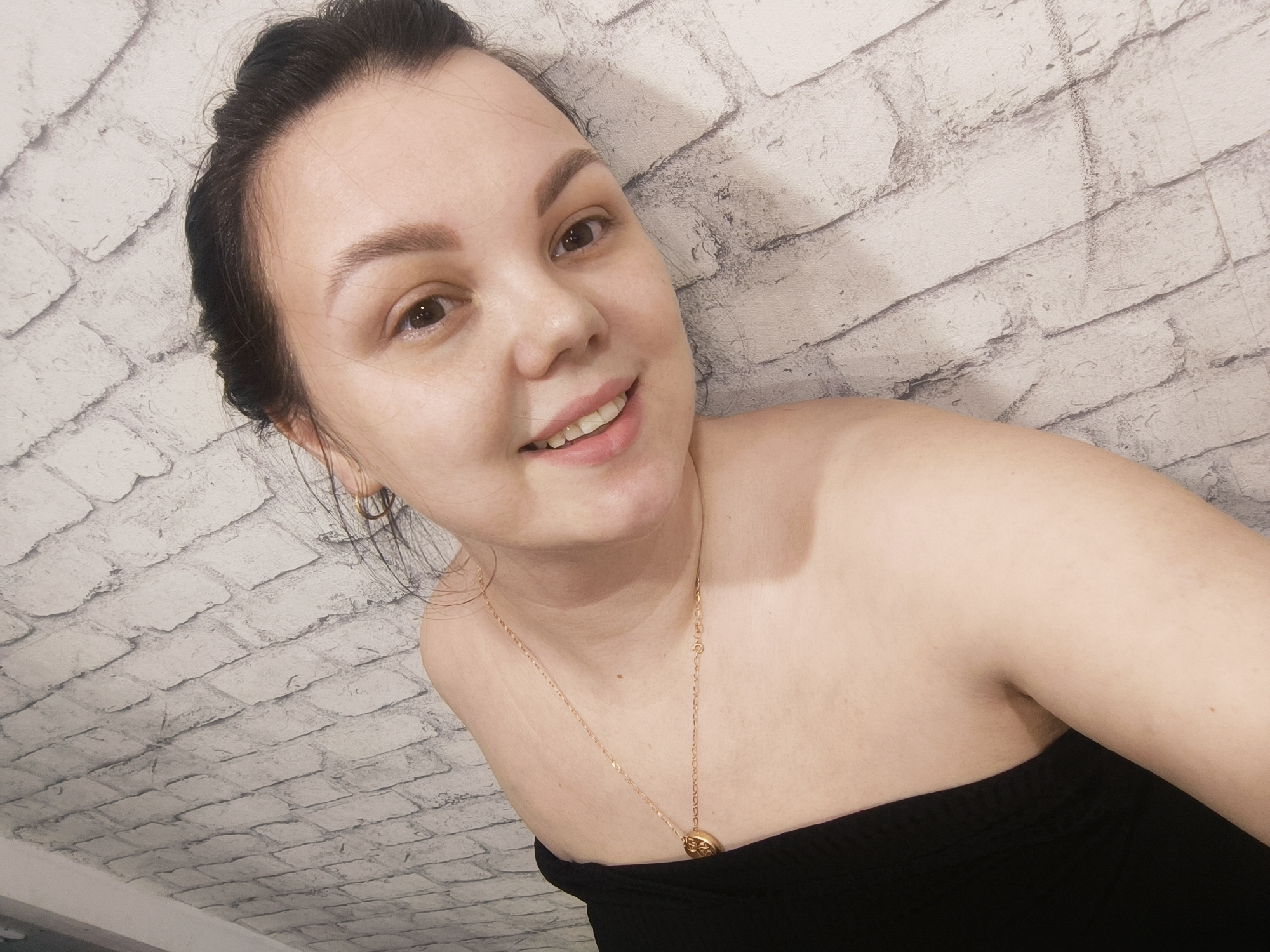 A woman with her hair pulled back is smiling and looking at the camera. She is wearing a black strapless top and a gold necklace with a pendant. She is positioned against a white brick wall. The location is Unknown, Unknown.