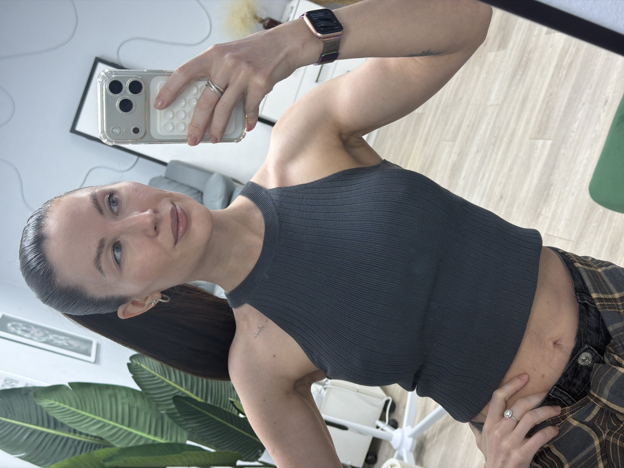 A woman is taking a selfie in a room with light-colored, wood-look flooring. She is holding a smartphone in her left hand, and her right hand is resting on her hip. She is wearing a dark, ribbed tank top and plaid pants. Her hair is pulled back. Visible on her left wrist is a smartwatch with a metallic band. In the background, there is a green plant, a framed artwork on the wall, a white office chair, and part of a gray couch. The location is not identifiable as Perm, Russia, based on the visual information provided.