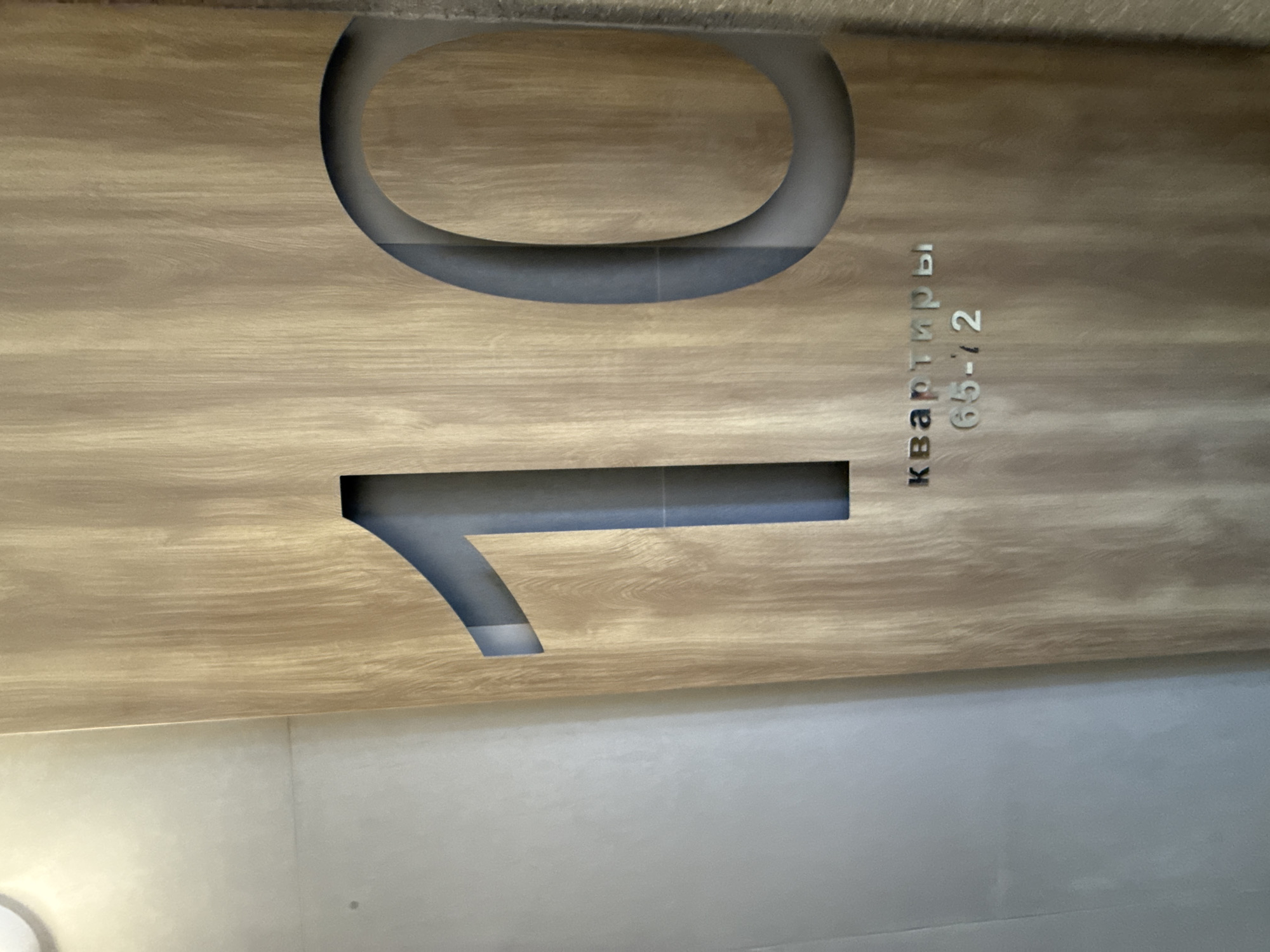 The image shows a sign indicating apartments 65-72 on the 10th floor. The background is light wood-grained and the lettering is gray. The location, Ovoshchnoy, Russia, is based on the prompt, not the visual evidence.
