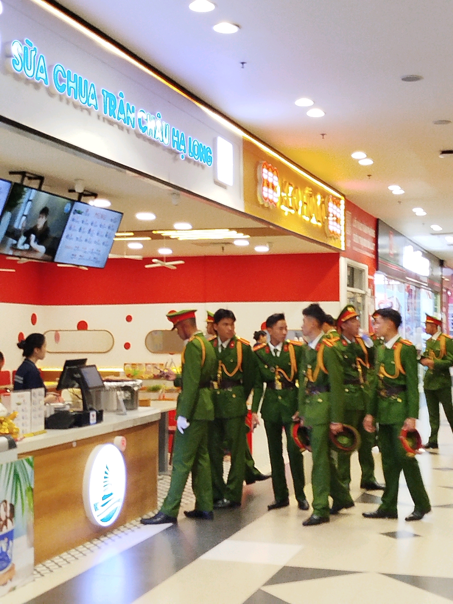 This is a medium shot of a group of Vietnamese soldiers in green uniforms and red hats standing in a line inside a brightly lit indoor market or shopping mall. Some of the soldiers are holding their hats in their hands. In the background, there is a red and white wall, a counter with food and drinks on display, and a large screen showing images. The soldiers appear to be waiting or queuing for something. The text on the blue neon sign above the soldiers reads "SỮA CHUA TRÂN CHÂU HẠ LONG". The floor is tiled with a geometric pattern of white and grey. The overall atmosphere is one of order and uniformity. The image does not provide any specific information to indicate the location in the city of Da Lat, Vietnam.