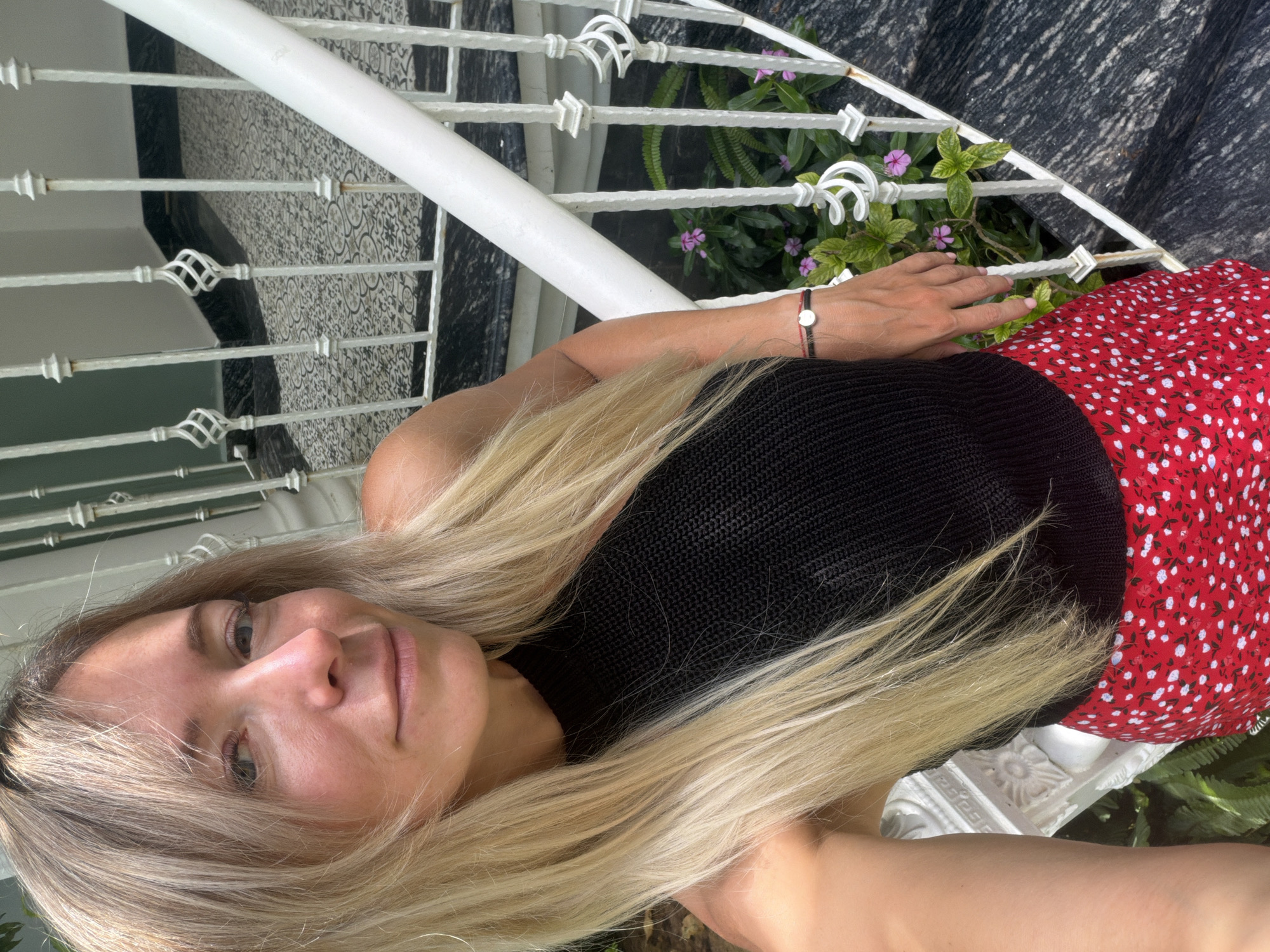 A young Caucasian woman with blonde hair and fair skin is posing for a selfie on an outdoor staircase. She is wearing a black sleeveless top and a red skirt with white polka dots. Her right arm is resting on a white railing, and she is wearing a bracelet on her wrist. Behind her, a white metal railing with decorative swirls lines the staircase, and there are green plants with small purple flowers growing along the side. The staircase has dark stone steps with a patterned tile runner. The overall lighting suggests it is daytime, with sunlight casting shadows. The location is identified as a staircase in the city of Xuân Trường - Đà Lạt, Vietnam.