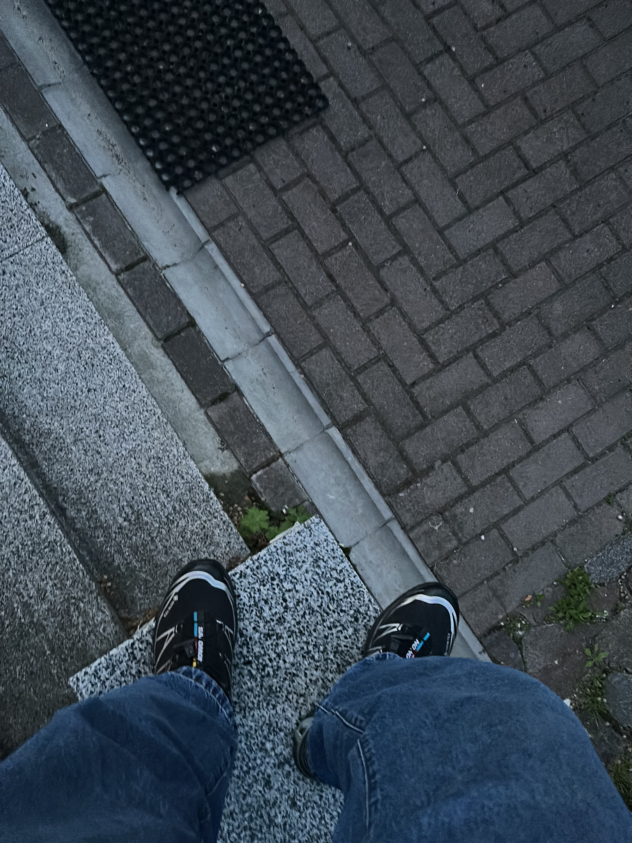 A first-person perspective photograph shows a person standing on a grey granite step, likely an exterior step of a building or entrance. The person is wearing blue jeans and black Salomon hiking shoes. To the right of the step is a pavement made of grey brick arranged in a herringbone pattern. A grey concrete curb separates the step from the brick pavement. A black rubber mat is on the brick pavement in the upper left corner of the image. Small plants can be seen growing between the step and the curb. The location is Kriukivshchyna, Ukraine.