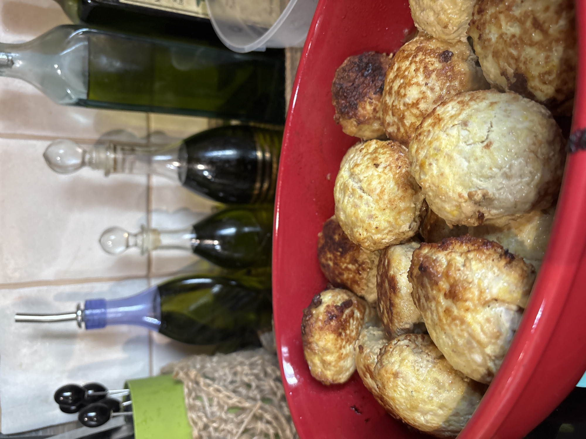 The image shows a close-up view of cooked meatballs in a red bowl, placed to the right of several green glass bottles. The meatballs are golden brown with some darker, slightly charred edges, indicating they have been pan-fried or baked. They are roughly spherical in shape and piled together in the bowl.
To the left of the bowl, several green glass bottles are arranged vertically. One bottle in the foreground has a purple spout with a silver tip, suggesting it might contain oil or vinegar. Below this bottle, a pair of black olive-shaped objects with white stems are visible, possibly part of a utensil set or food decoration. The bottles appear to be on a tiled surface, with a textured material, possibly burlap or twine, visible at the bottom left of the image.
The lighting suggests an indoor setting, possibly a kitchen or dining area. There is no indication of the time of day or weather conditions. The image is a still photograph and does not depict any specific event or activity beyond the presentation of the food and bottles. No visible text is present in the image. The overall impression is one of food preparation or serving.