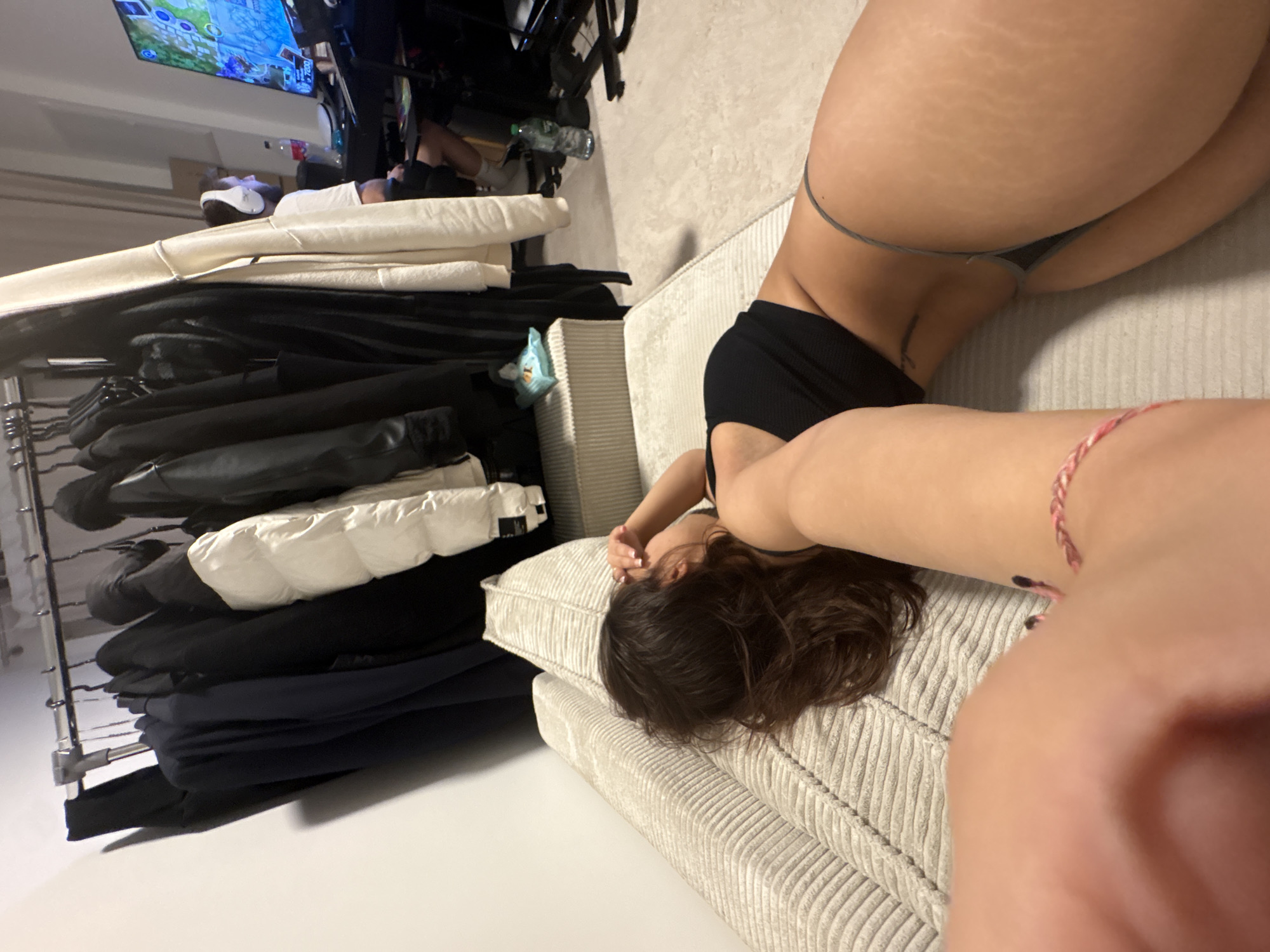 The image shows a woman lying on a light-colored, textured sofa. She is wearing a black top and a thong. A rack of clothing, including jackets and coats, is visible on the left side of the frame. In the background, a person is wearing a VR headset and playing a game on a screen. The location is Mönchengladbach, Germany.