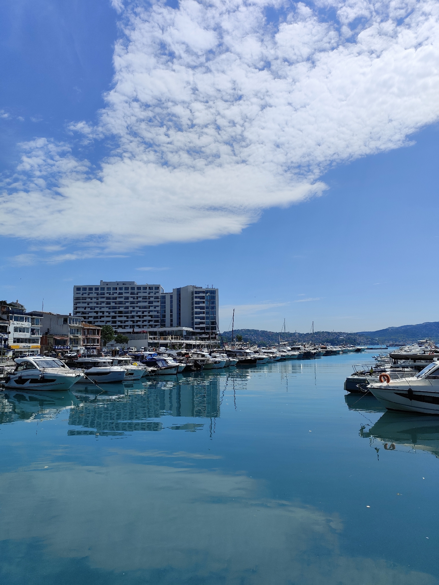 This media file captures a picturesque marina scene in Sarıyer, Turkey, under a clear, bright blue sky dotted with scattered white cumulus clouds. The image is dominated by the calm, turquoise-blue water of the marina, which acts as a mirror, reflecting the sky's patterns and the buildings lining the shore.
Numerous modern boats and yachts of various sizes are moored neatly along the docks, stretching into the distance. Their sleek white hulls and polished surfaces create a sense of luxury and leisure.
Along the waterfront, a prominent, multi-story modern building with a facade of many windows stands tall in the background, possibly a hotel or apartment complex. To its left, a series of smaller, multi-level buildings with balconies and terraces line the shore, appearing to house various businesses or residences. Visible text on one of these buildings includes "FILIZO..." and "İyilik Bankası." Another sign partially shows the word "LİFE." In the far distance, gentle green hills or mountains frame the horizon under the vast sky.
The scene is bathed in bright daylight, suggesting a sunny morning or early afternoon. The overall atmosphere is tranquil and serene, with no discernible human activity or people in view. The clear reflections on the water emphasize the calm conditions and the beauty of the coastal setting.