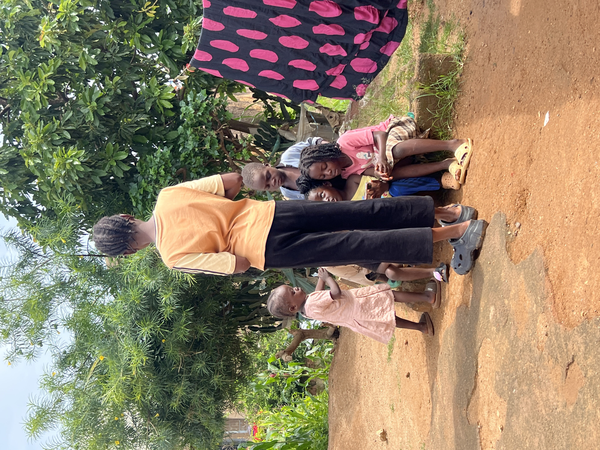 An outdoor scene in Jos, Nigeria, depicts a group of children and an older youth under the shade of lush green trees. The ground is composed of reddish-brown earth, with some paved areas. A black and pink patterned cloth hangs to the right, possibly drying laundry.
In the foreground, a young girl in a light pink dress stands, looking towards the left. To her right, a group of three younger children are seated. One child, wearing a pink t-shirt and a checkered skirt, is looking down. Another child, wearing a light blue top, is looking forward. A third child, whose face is mostly obscured, is also seated with the group.
Behind the seated children, an older youth, seen from the back, is wearing an orange t-shirt with yellow stripes on the sleeves and black pants. The youth's braided hair is visible.
The environment suggests a residential area with abundant vegetation, including corn stalks visible in the background to the left. The lighting indicates it is daytime.