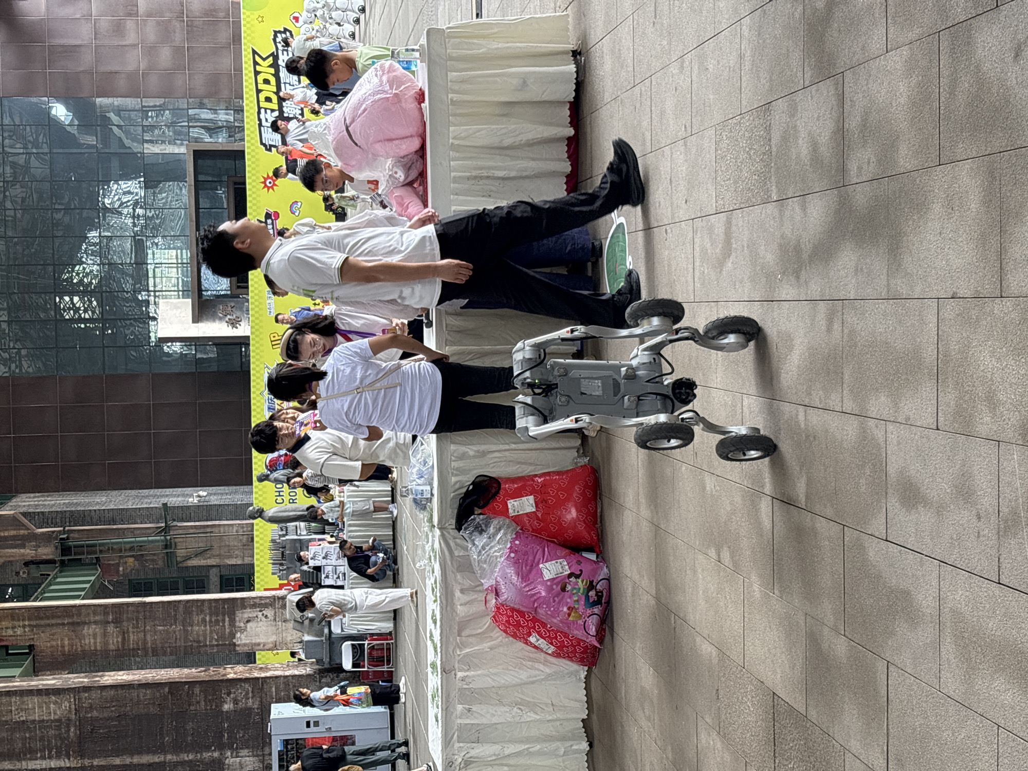 In Dadukou, China, a group of people stand around a table covered with a white cloth, presenting various items. A robot with two wheels in the front and two in the back is positioned in the foreground, facing away from the viewer. Three large, bagged items, one pink with cartoon characters and two red with heart patterns, are placed on the ground in front of the table. Behind the table, several individuals, including men, women, and children, are visible. The background features a large, modern building with a glass facade. The ground is paved with square tiles.
