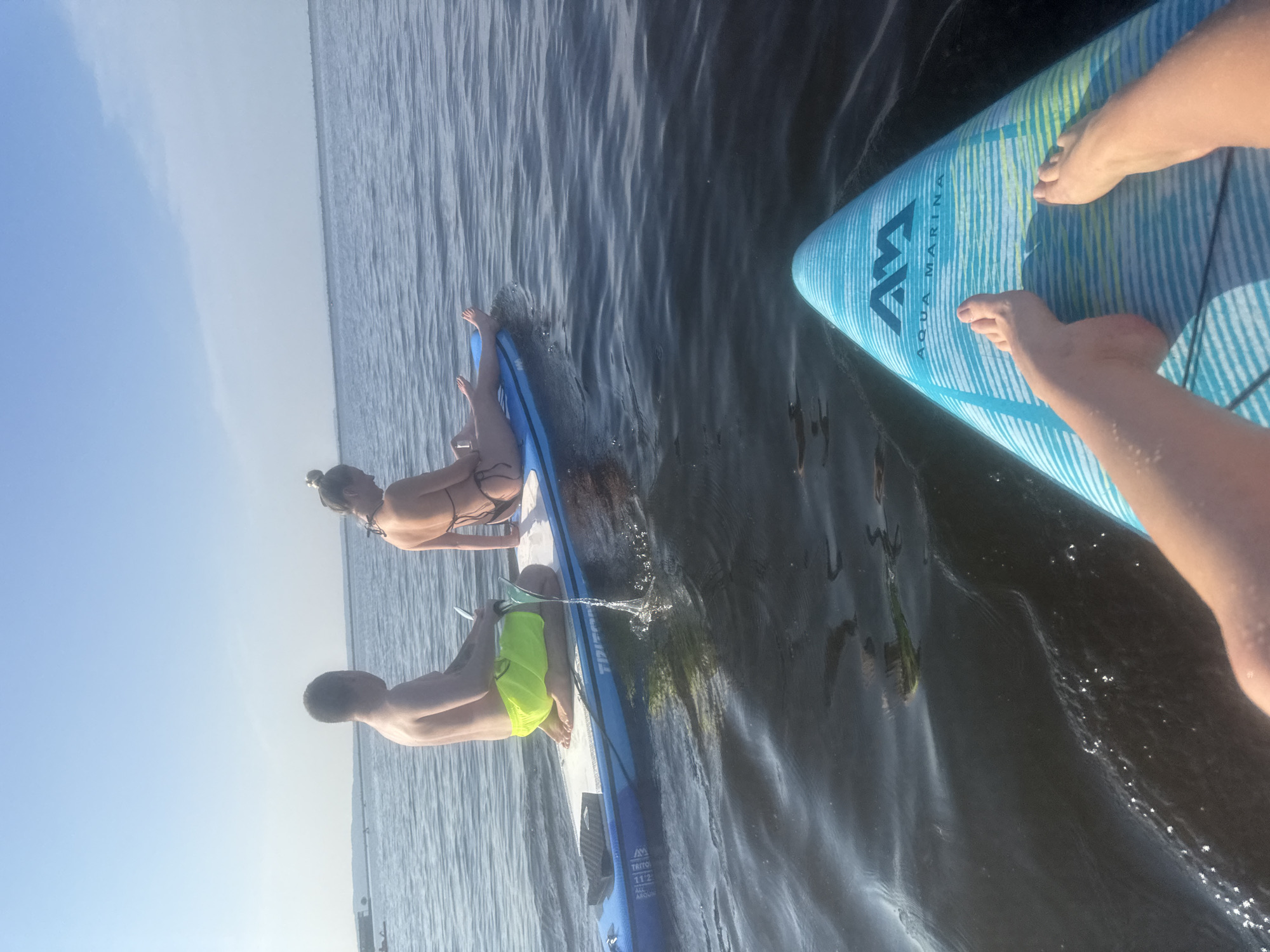In Chaikovsky, Russia, two individuals are paddleboarding on a body of water under a clear sky. A man in neon yellow shorts sits facing away from the camera, holding a paddle. A woman in a black bikini sits facing the same direction. In the foreground, a person's feet are visible on another paddleboard. The water is dark and rippled, reflecting the sky. The scene is captured from a low angle, looking across the water.