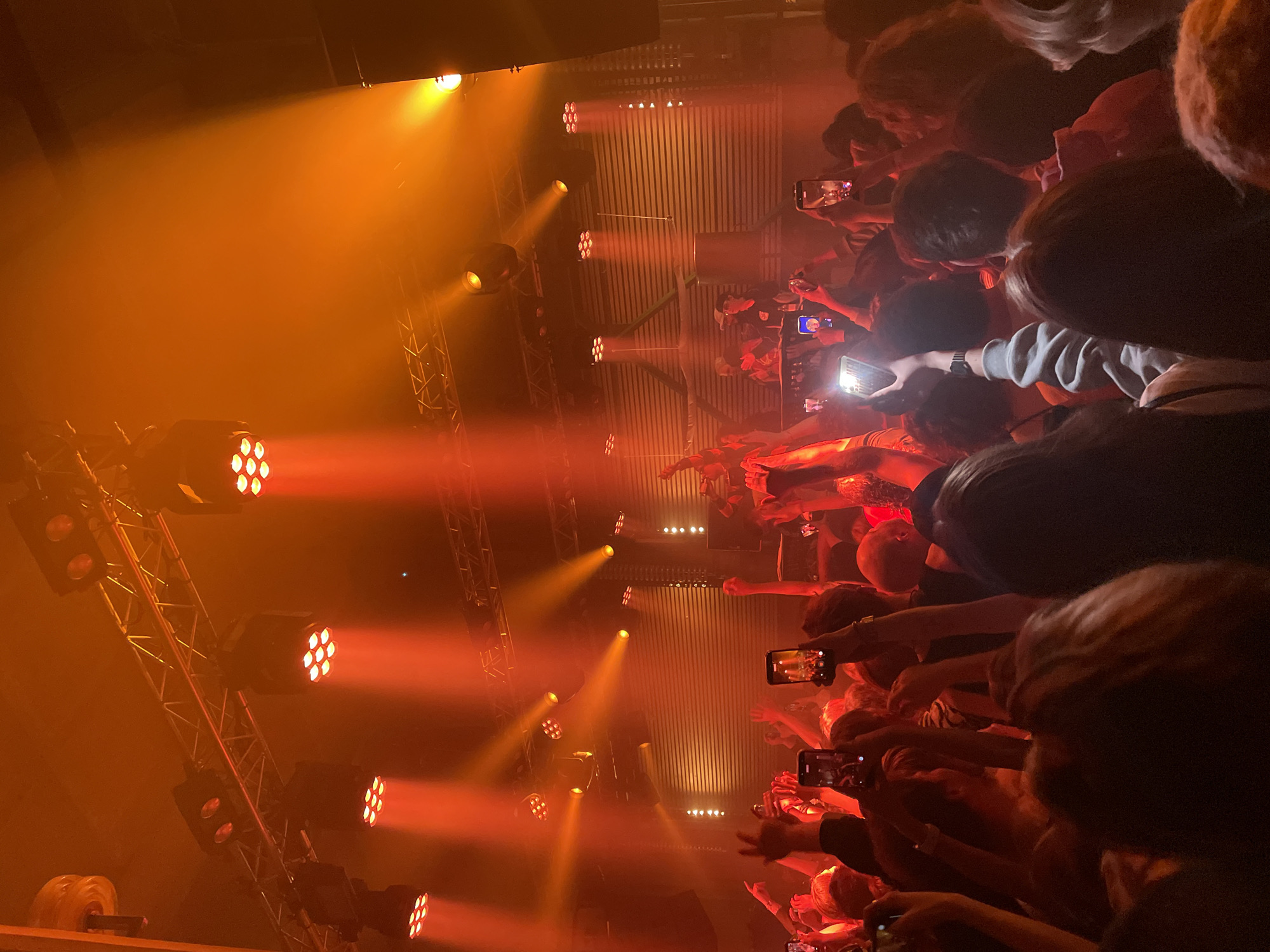 The image depicts a concert or performance event, likely an indoor venue, as evidenced by the stage lighting rig on the left side of the frame. The location is Saint Petersburg, Russia. A crowd of people is visible from above, facing towards the stage. Many individuals are holding up smartphones, presumably to record or photograph the performance. The lighting consists of multiple spotlights that emit an orange glow, creating a hazy and smoky atmosphere. The scene is bathed in warm, intense light, typical of stage productions. The backdrop appears to be a corrugated metal wall behind the performance.