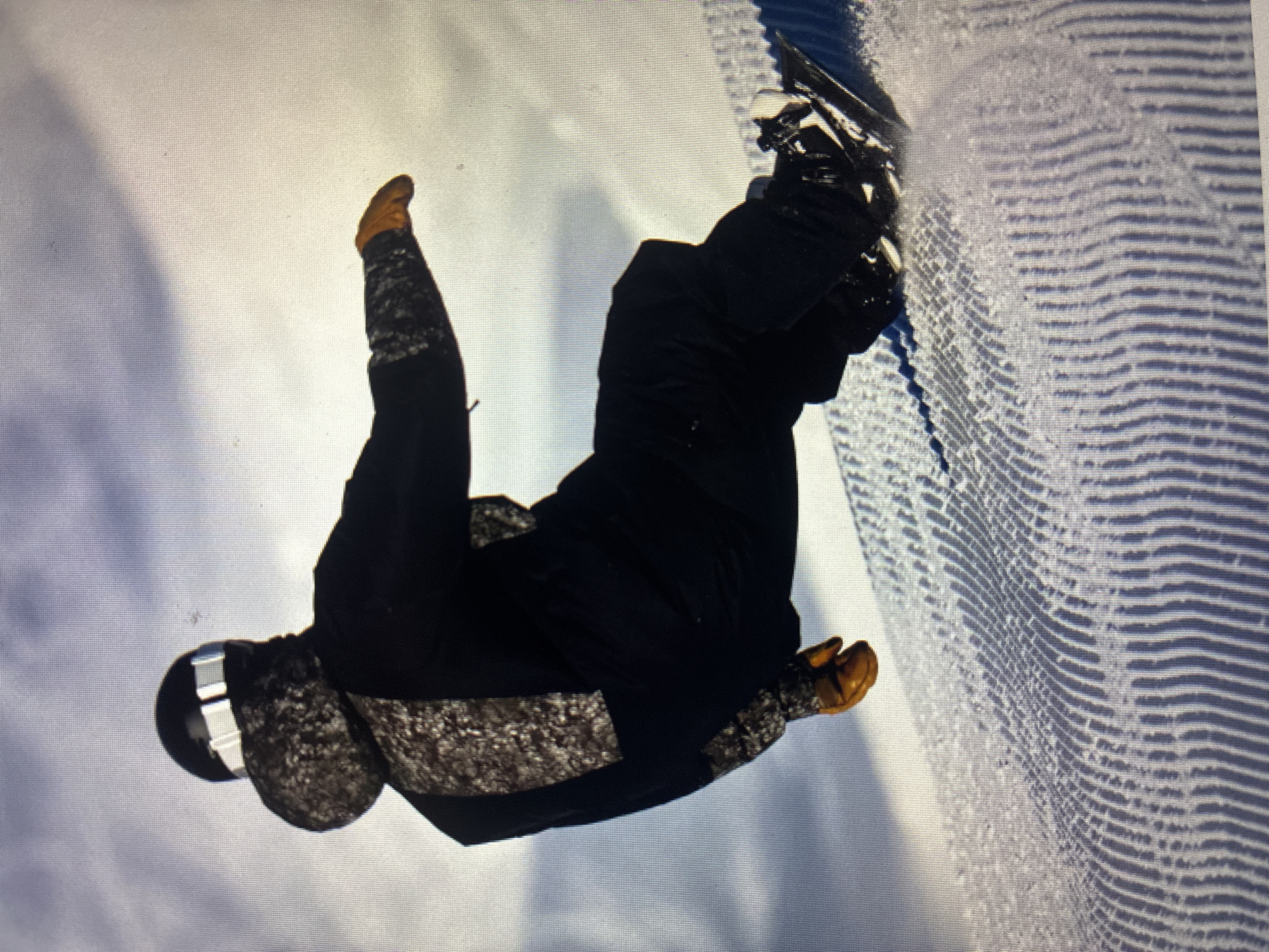 The image captures a snowboarder in mid-air or executing a deep carve on a snow-covered slope, with the photograph rotated 90 degrees clockwise. The snowboarder is dressed in dark winter apparel, including a black jacket and pants, featuring distinct patterned sections on the lower sleeves and potentially around the headwear, possibly a helmet. Light brown or tan gloves are visible on the hands, and a dark helmet with a reflective silver or white accent covers the head. A dark snowboard is clearly visible, attached to the rider's feet by bindings, suggesting dynamic movement as evidenced by light snow spray around the board.
The setting is a brightly lit, snowy environment, likely a groomed snow park or half-pipe. Prominent parallel lines from snow grooming equipment are etched into the snow, particularly on what appears to be the wall or ground of the snow feature on the right side of the image. A thin blue line or stripe is also visible along the edge of the snow surface. The overall impression is one of action and skilled athleticism.
The scene takes place during the daytime under bright illumination. No specific weather conditions are discernible beyond the presence of snow, and visibility appears clear. There is no visible text within the image. The provided location context indicates this activity occurred in Asenovgrad, Bulgaria.