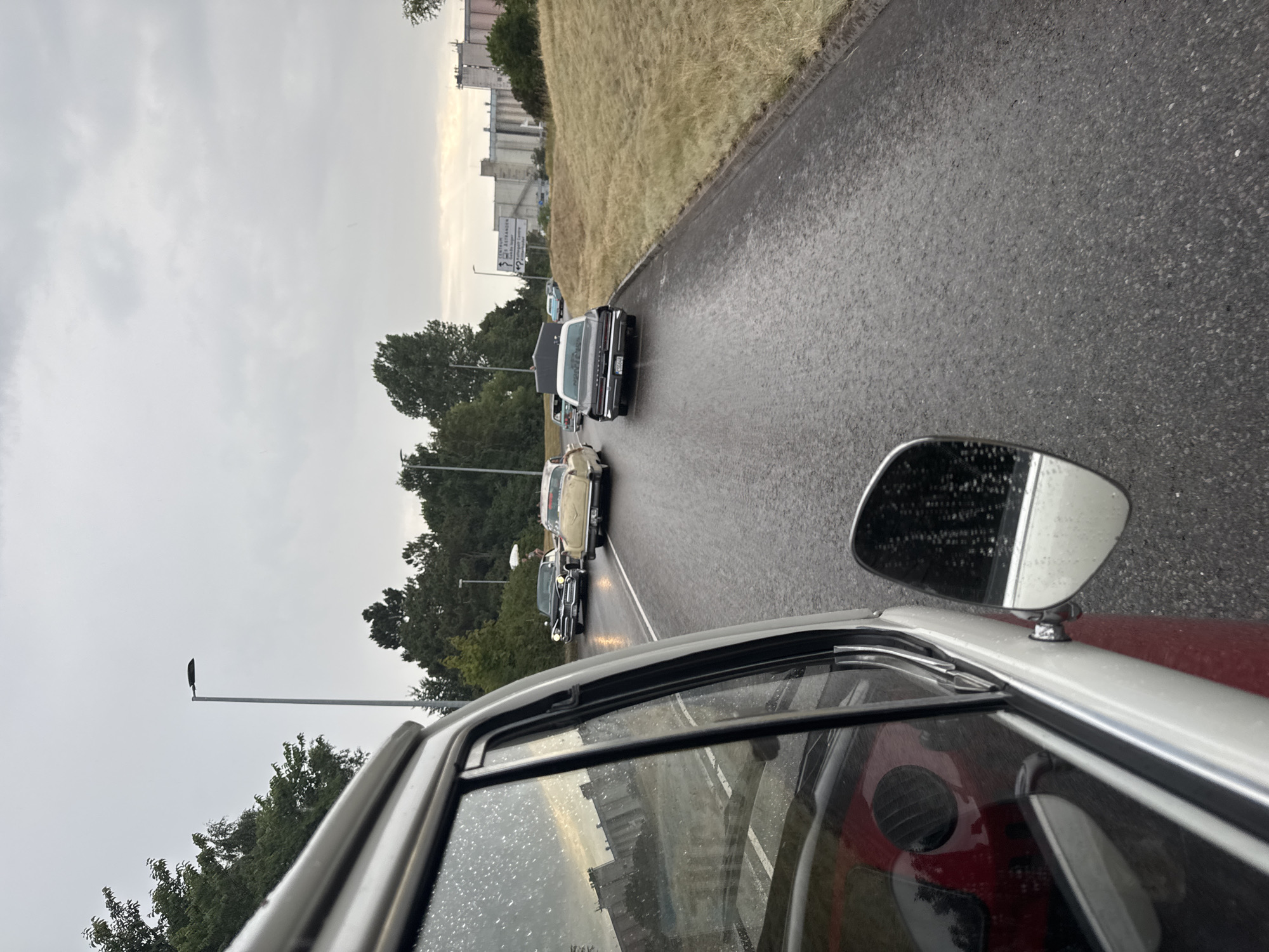 The image captures a convoy of vintage cars driving on a wet asphalt road in Falkenberg, Sweden, under an overcast sky. The perspective is from inside one of the vehicles, looking forward. The foreground is dominated by the interior of the car, including a portion of the dashboard and window, with raindrops visible on the glass. Several classic cars, including a black sedan with a white roof and a cream-colored sedan, are visible ahead on the road. The road is lined with dry, yellowed grass and some trees. In the background, industrial buildings and a signpost indicating "CENTRUM ÅSTRANDEN" and "Falkenbergs Centrum" can be seen. The overall scene suggests a car meet or parade taking place in potentially inclement weather.