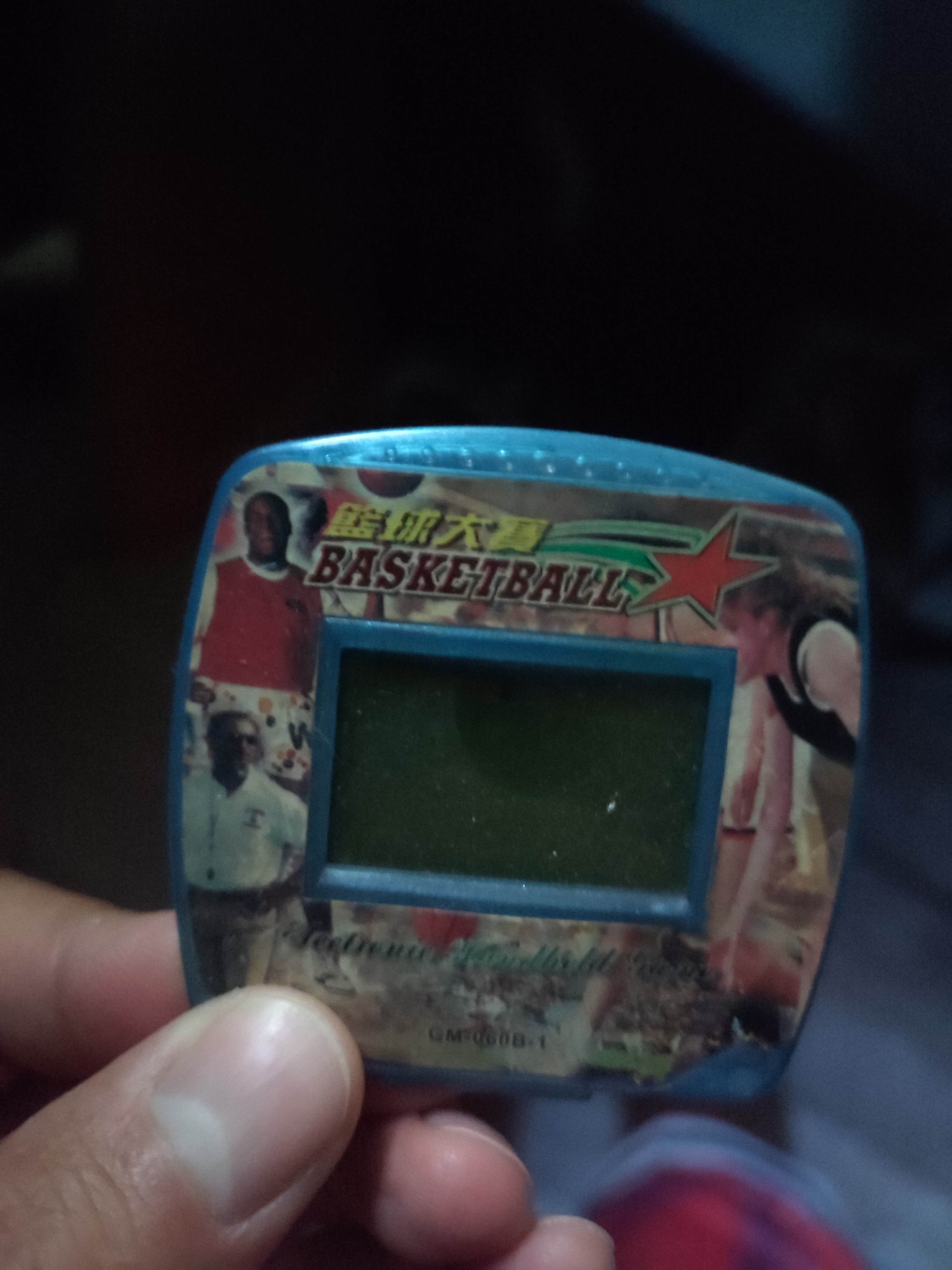 An unidentified hand holds a small, blue, rectangular electronic toy. The toy has a screen in the center and is decorated with images of basketball players and the word "BASKETBALL" in English, along with Chinese characters and a red star. The background is dark and out of focus. The toy appears to be an old handheld electronic game. There is no information to indicate this image is located in Gremyachinsk, Russia.