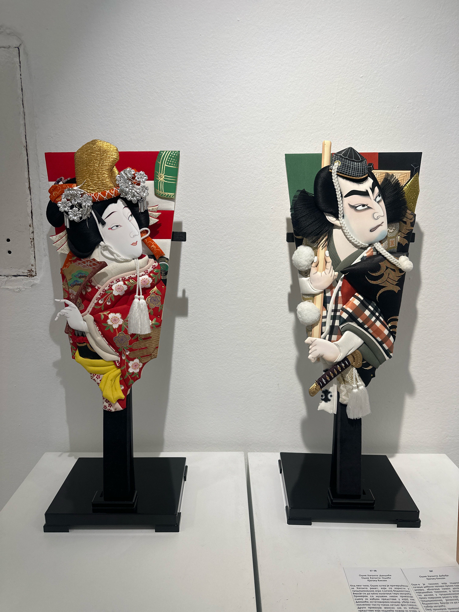 The image shows two traditional Japanese doll figures displayed on black pedestals on a white surface, likely in a museum or gallery. The figure on the left depicts a geisha or a female kabuki actor, adorned in a vibrant red kimono with floral patterns, elaborate gold headdress, and ornate silver hair accessories. The figure on the right represents a male kabuki actor, characterized by his dramatic makeup, black wig, and a kimono with a checked and patterned design, holding what appears to be a fan and a sword. Both figures are mounted on a fan-shaped backdrop featuring a mix of colors and patterns, including red, green, black, and gold. The background is a white wall with some subtle texture and a dark mark on the left. There is a printed label with Cyrillic text visible on the white surface in the lower right corner, indicating the location is in Belgrade, Serbia.