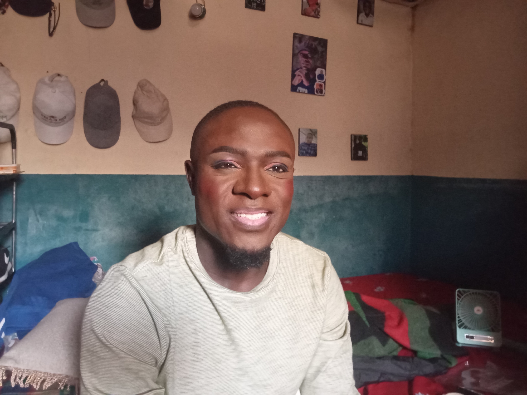 A person with dark skin, wearing makeup including eyeshadow and blush, is pictured in a room with a textured, pale yellow wall. The person is wearing a light green, striped t-shirt. Several caps are displayed on the wall behind them, including white, grey, and beige ones. Small framed pictures of people are also visible on the wall. In the foreground, a blue blanket and a beige cushion are present. A small, light blue fan sits on a red surface on the right side of the image. The overall impression is of an indoor setting.