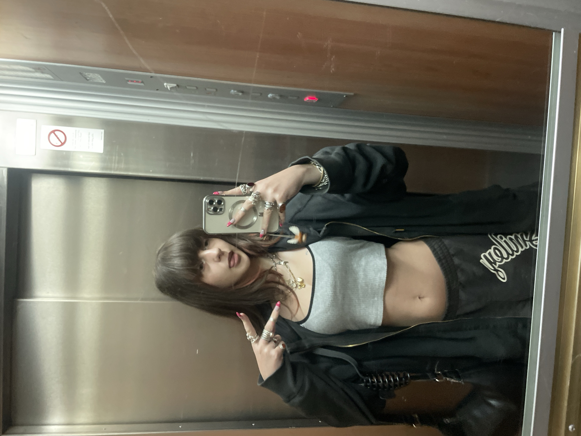 A female individual is depicted taking a mirror selfie inside an elevator in Petah Tikva, Israel. She is centrally positioned, facing her reflection and holding a smartphone with a multi-lens camera array in her right hand to capture the image. Her left hand is raised, forming a peace sign. She wears multiple silver-toned rings on both hands, some with visible red or dark stones, and her fingernails are painted bright pink. She is dressed in a light grey ribbed crop top, an unzipped dark hoodie, and dark sweatpants with a white script logo on the left thigh. Her midriff and navel are exposed. Several silver-toned necklaces with various pendants are visible around her neck.
The elevator interior features polished metal or mirrored surfaces on the left, consistent with brushed stainless steel doors, and a wood-patterned or brown panel on the right. A grey elevator control panel is affixed to the right wall, displaying several buttons, one of which is illuminated red. A white rectangular sign with a red circular 'no entry' symbol and text in Hebrew and English is visible on the elevator door above the subject's head. The overall lighting is bright, illuminating the reflective surfaces.