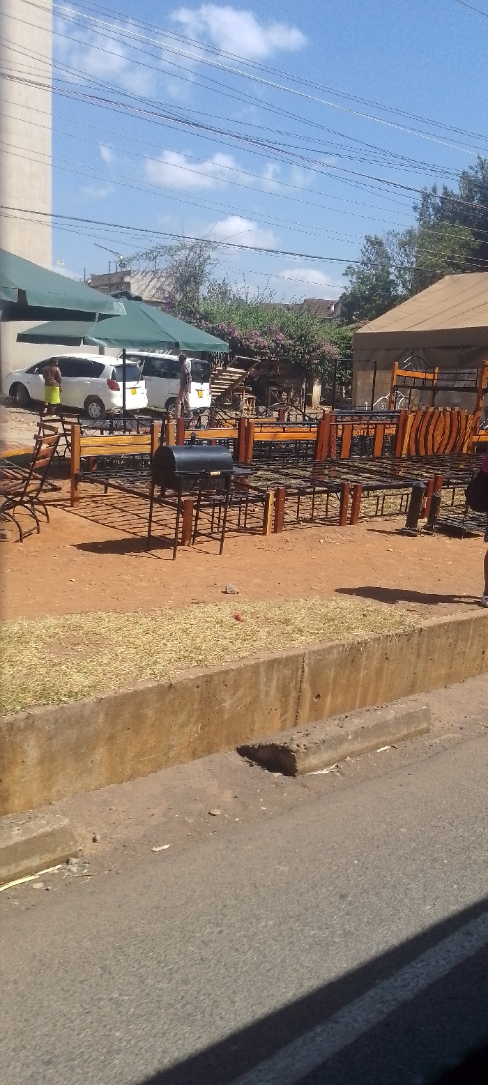 The image captures an outdoor commercial scene in Ngong ward, Kenya, under a clear blue sky with scattered white clouds and numerous overhead power lines. The foreground features a paved asphalt road separated by a concrete curb from a narrow strip of dried grass, which then transitions into a reddish-brown dirt area. This dirt area displays various metal bed frames, some incorporating wooden headboards or footboards, arranged in rows. A black, lidded barbecue grill on a stand is centrally positioned among the bed frames.
To the left, two white vehicles, identified as a hatchback and a van, are parked beneath two large green awnings. A person wearing a light top and a green skirt stands near the left white vehicle. Another person is partially visible near the white van. On the far right, only the lower leg and white shoe of a person are visible, standing near the displayed bed frames. Behind the vehicles and awnings, a light-colored building is visible on the left, and a structure with a brown, tent-like roof is present on the right. The background features green trees, flowering bushes, and additional distant buildings. Shadows indicate direct sunlight.