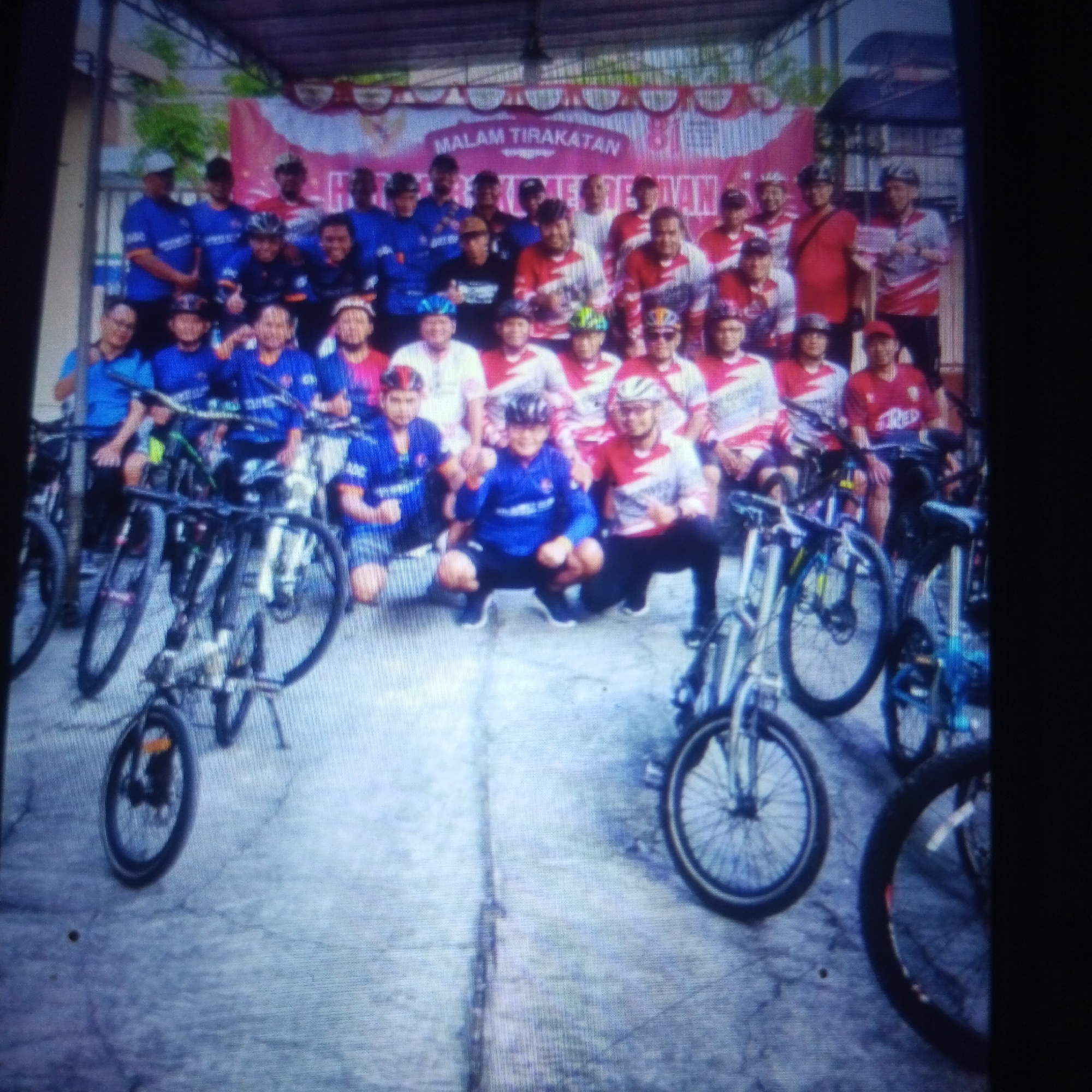 A group of at least 20 individuals, all appearing to be adult males, are posing for a photograph outdoors in front of a banner. The banner, with red and white decorative elements, reads "MALAM TIRAKATAN" and appears to be in Indonesian. The individuals are wearing athletic clothing, mostly t-shirts and shorts, with many sporting cycling helmets. Several bicycles are visible in the foreground and to the sides, suggesting a cycling event or gathering. The ground is paved and appears to be a concrete or asphalt surface, with some areas of wear and tear. The overall setting suggests a community event, possibly related to cycling, taking place in an outdoor public space in Cemani, Indonesia.