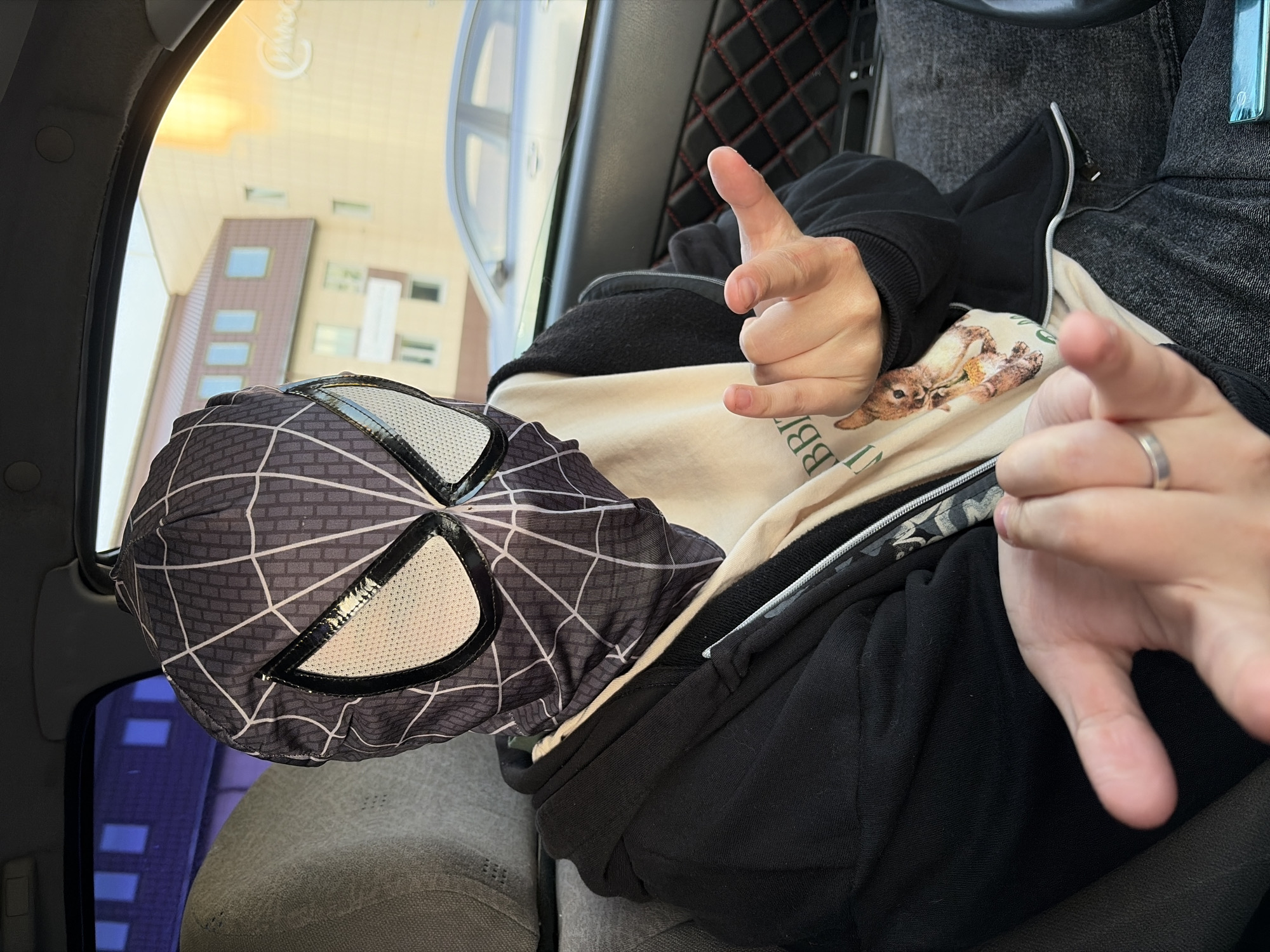 The image, rotated 90 degrees counter-clockwise, depicts a person inside a car in Chaikovsky, Russia, during daytime. The individual, likely a young adult, is seated, wearing a black zip-up hoodie over a light-colored sweatshirt featuring a graphic of a small, light-brown mammal (resembling a squirrel or marmot) holding food. A black and grey Spider-Man mask, designed after the black suit, rests on their lap or the car seat.
The person's hands are prominent, making two distinct gestures. The hand closer to the viewer (subject's left hand) displays the index finger and pinky extended, along with the thumb, while the middle two fingers are curled, forming a "rock on" or "shaka" sign. The other hand (subject's right hand) shows the index finger and thumb extended with the remaining fingers curled, typically used for pointing or as a "call me" gesture. A silver ring is visible on the ring finger of this hand.
Through the car windows, multiple buildings with modern architecture and large rectangular windows are visible under a bright, clear sky. The buildings are primarily light brown or tan. The car's interior features dark upholstery, including a diamond-quilted pattern on the back of the seat. Faint, reversed text reading "Midea" is visible as a reflection on a window, and partial text, possibly "BBIR" or "BBIS", can be seen on the subject's sweatshirt.