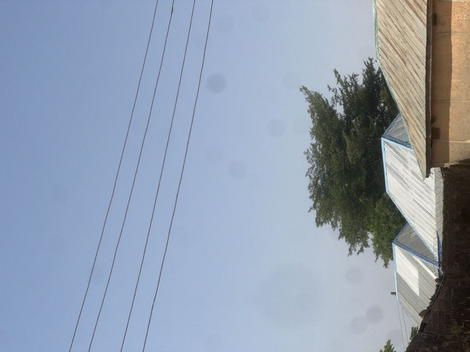 The image displays an outdoor scene in what is identified as a residential area of Birnin Kebbi, Nigeria, under a pale blue sky. The upper third of the frame shows a clear sky horizontally traversed by four parallel power lines. Scattered across the sky are multiple faint, dark, circular artifacts, consistent with lens dust or sensor smudges. Below the power lines, a large, dense, dark green tree occupies the central portion of the mid-ground. In the foreground, the upper sections of multiple buildings are visible. Two prominent structures feature sharply angled, corrugated metal roofs; these roofs are silver in color with light blue trim along their edges. To the immediate right, another building's flatter roof, constructed from rusted, brown corrugated metal, is partially visible, with a section of its light brown or ochre-colored wall beneath it. A dark, rough-textured wall, likely constructed from brick or concrete blocks, runs across the lower left foreground, obscuring the base of the structures and the lower part of the tree. No individuals or active movements are depicted in the static scene.