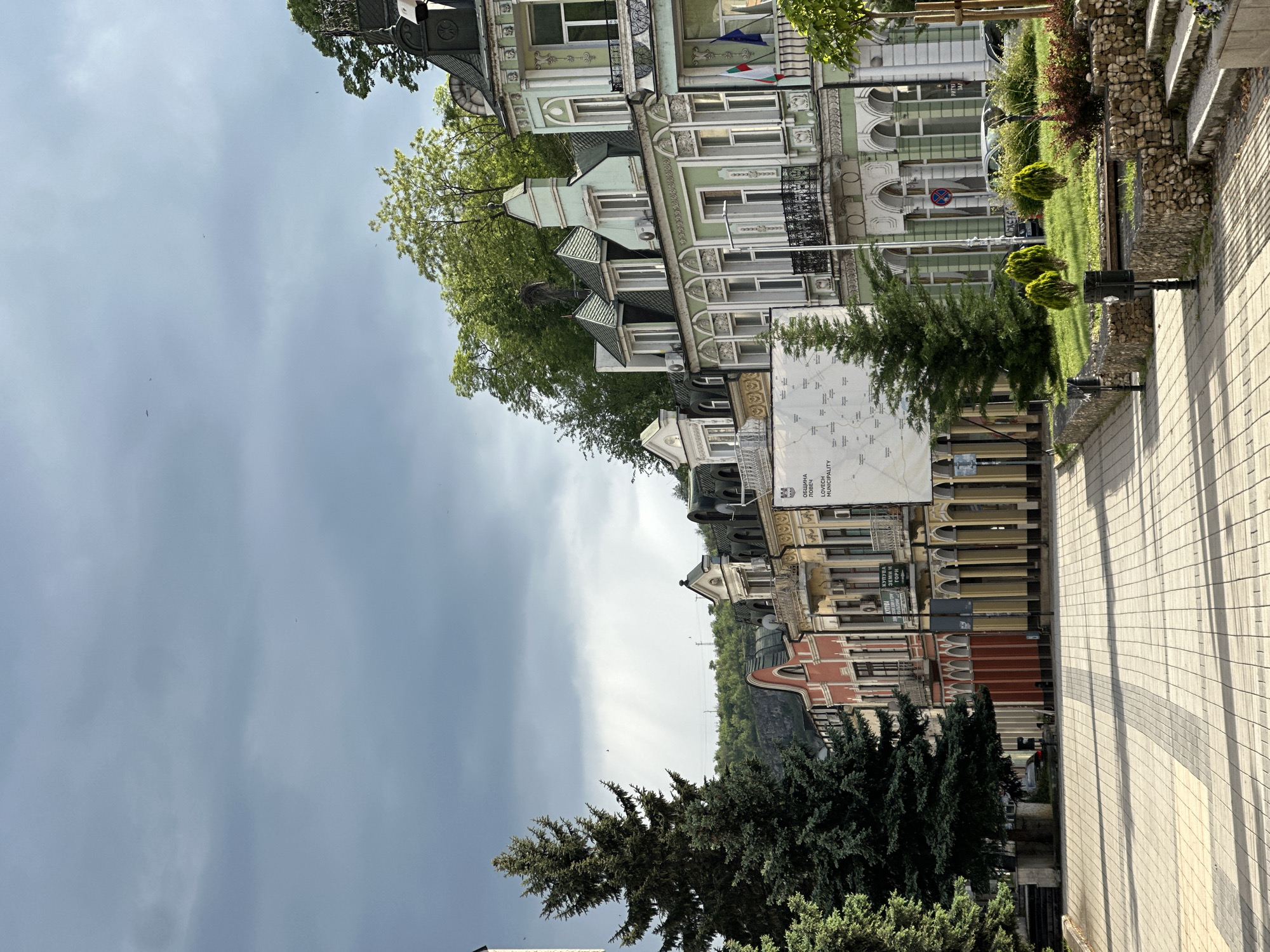 The image shows a street scene in Lovech, Bulgaria. Ornate, historical buildings line the street, some with green and white facades. A map of the Lovech Municipality is displayed on one building. The street is paved with rectangular blocks and lined with trees and small shrubs. The sky is overcast with dark clouds. No people are visible.