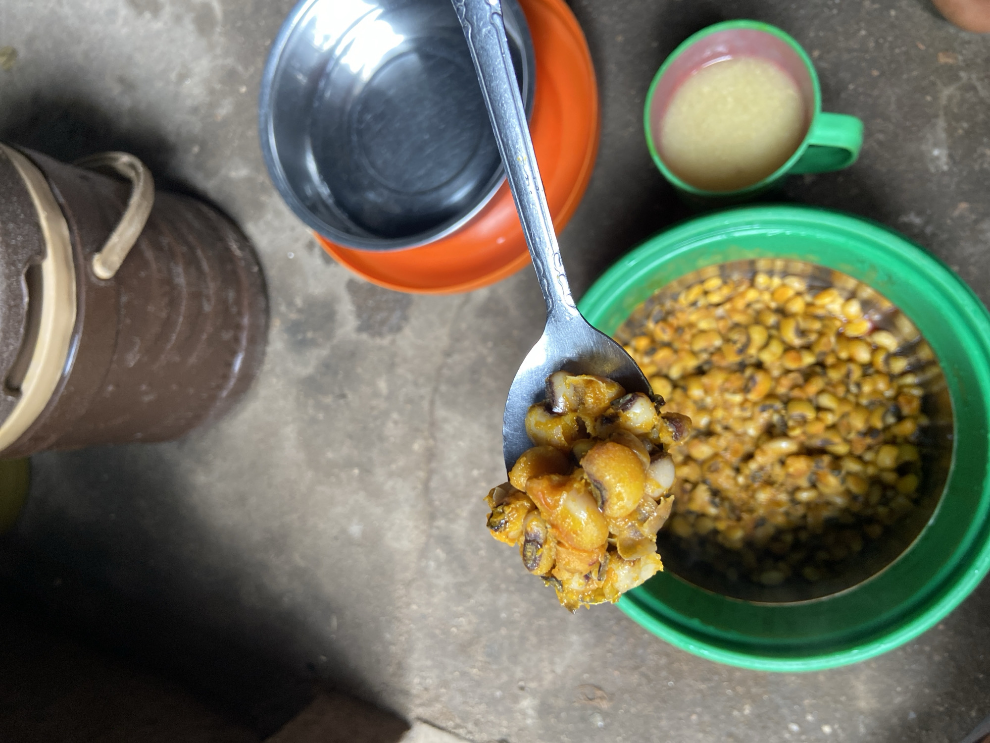 A close-up, high-angle shot captures a spoonful of cooked black-eyed peas being lifted from a green plastic bowl filled with the same dish. The peas are coated in a yellow-orange spice mixture. To the right, a small green plastic cup holds a pale yellow liquid. Behind the bowl, an orange plastic plate supports a shiny metal bowl. In the background, a large brown plastic container with a tan lid and handle sits on a concrete floor. The scene appears to be set in an outdoor or semi-outdoor environment in Mararaba, Nigeria, given the rustic concrete flooring and the type of food preparation and serving items visible.