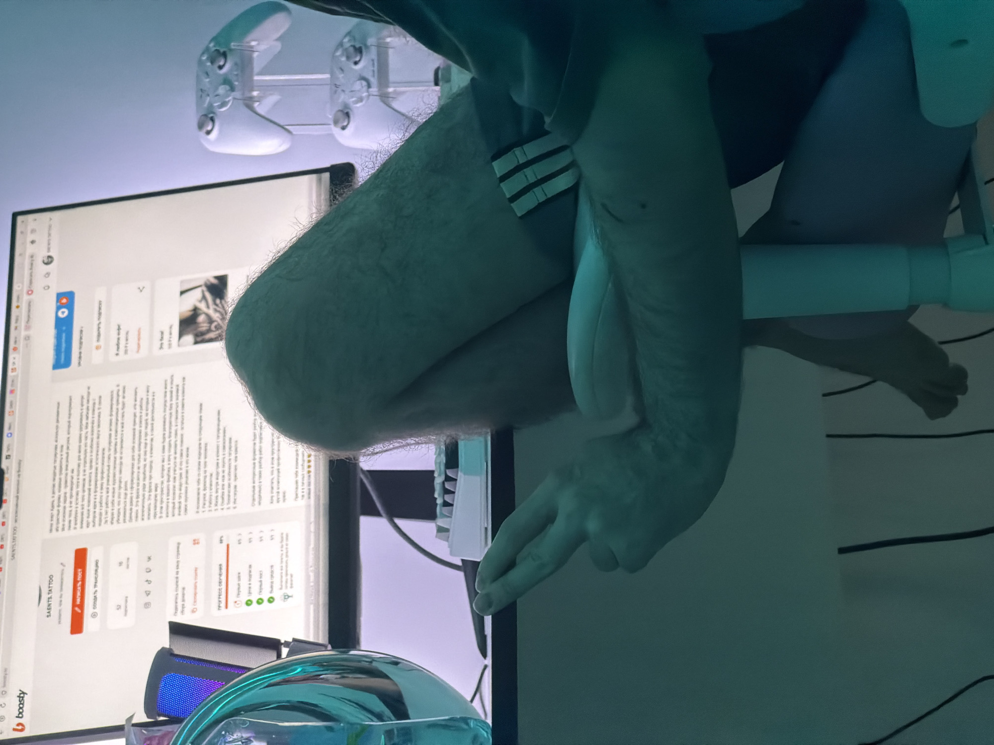 This indoor photograph, taken in Khimki, Russia, captures a person seated at a desk, interacting with a computer. The image is taken from a low angle, showing the person's legs, one arm, and hand in the foreground, with a monitor, gaming controller, and other desk items in the background.
The visible subject is a person, likely male, wearing shorts that reveal hairy legs. A white band with three black stripes is visible on one thigh, possibly part of athletic wear or an accessory. One hand is positioned over what appears to be a keyboard or mouse area, with the index and middle fingers extended. The person's posture suggests engagement with the computer setup.
In the background, a large computer monitor displays a website with Russian text. The website appears to be "boosty.ru", a content creation platform, indicated by the "boosty" logo and the text "SAENTS TATTOO" as a page title or username. Visible elements on the screen include buttons like "Написать Пост" (Write a post) and "СООДАТЬ ТРАНсляцию" (Create a broadcast/stream), along with sections showing engagement numbers (e.g., 52, 16, 453, 10) and a "ПРОГРЕСС ОБУЧЕН" (Learning progress) status. Articles or posts with headlines like "Я люблю кофе!" (I love coffee!) and "Это весна!" (It's spring!) are also partly visible.
Above the monitor, a white gaming controller is mounted on a stand, possibly against a wall or shelf. A desk or surface below the monitor holds what appears to be a keyboard with white keys and a device emitting a blue light, possibly a microphone or speaker. A large, clear, teal-colored object, possibly a glass or a decorative item, is seen in the bottom left foreground. The lighting in the room has a cool, greenish-blue tint, suggesting artificial illumination. The overall scene depicts a casual, modern workspace or gaming setup.