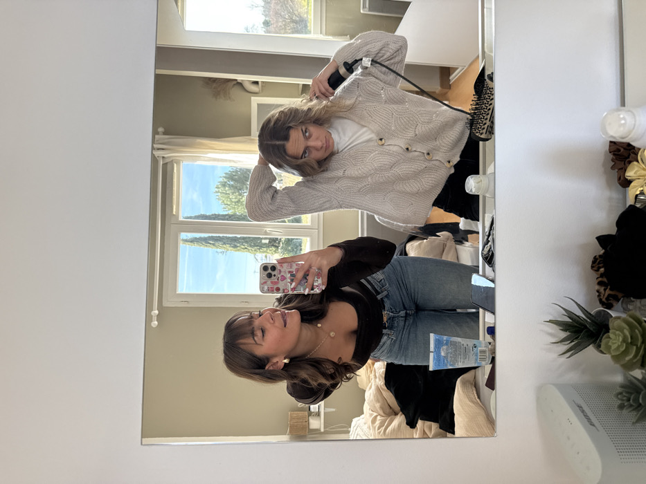 Two young adult females are reflected in a rectangular mirror, positioned inside a residence in Veauchette, France.
The female on the left, with brown hair and bangs, wears a dark brown long-sleeved top, blue jeans with a gold belt buckle, gold hoop earrings, and a gold pendant necklace. She holds a white iPhone with a colorful patterned case in her right hand, angled to capture the mirror reflection. Her eyes are partially closed, and she has a slight smile.
The female on the right, with wavy brown hair parted in the middle, wears a white turtleneck shirt underneath a light beige knit cardigan with a textured pattern and brown buttons, and dark trousers. She is actively styling her hair on the left side of her head with a black electric hair tool held in her right hand, while her left arm is raised. Her expression is focused.
On the white vanity surface in front of the mirror, from left to right, are a white Bose speaker, three small artificial succulent plants in pots, multiple hair scrunchies (including leopard print and solid colors), a small white dispenser bottle, a blue tube of product, a mobile phone lying flat, and a round hairbrush with a patterned handle.
The background reflection visible behind the individuals and through a large, white-framed window shows an interior room with light beige walls. A rumpled bed with light-colored bedding and dark clothing is visible on the left. Outside the window, a clear blue sky with sparse clouds is visible, along with two tall, slender evergreen trees (possibly cypress) and general green foliage, suggesting an outdoor natural setting. A white cabinet or wardrobe is visible on the far right of the reflection.
