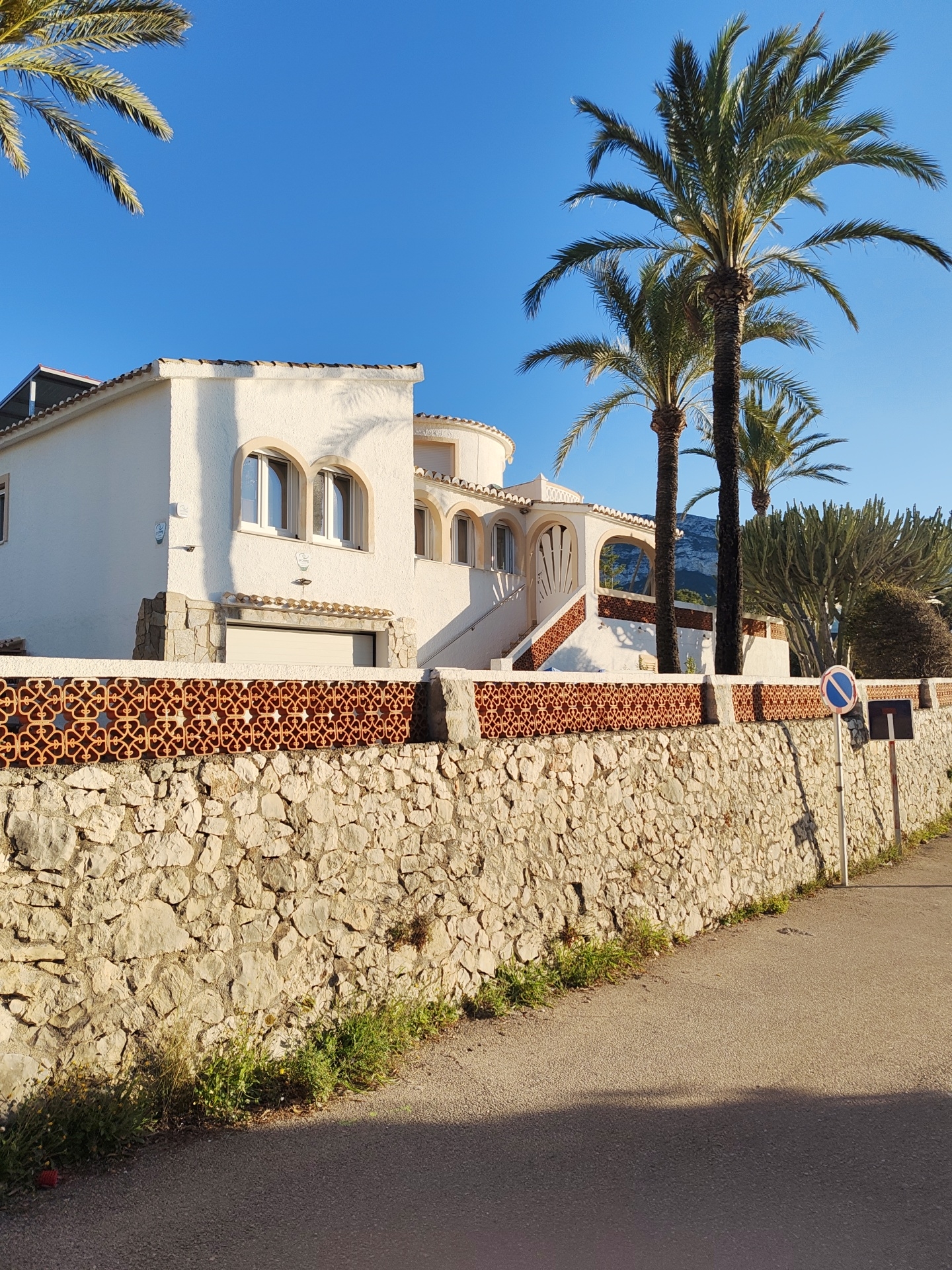 The image captures a vibrant, sunlit scene of a Mediterranean-style residence in Dénia, Spain, likely during the golden hour of late afternoon or early morning due to the warm light and long shadows. The main subject is a striking white house with terracotta roof tiles, featuring several arched windows and a prominent exterior staircase leading to an upper entrance. A rounded section of the house is visible on the right. Below the house and extending along the paved road, a robust stone wall with irregular blocks supports a distinctive reddish-brown terracotta lattice fence, showcasing intricate geometric patterns.
Multiple tall palm trees, characteristic of a warm climate, frame the house against a clear, deep blue sky. Another large, cactus-like plant with multiple branches stands to the right of the property. In the distant background, hazy mountains are visible, adding to the scenic setting. The asphalt road in the foreground shows a clear demarcation between sunlit and shadowed areas, with patches of green vegetation growing at its edge beside the stone wall. A "no stopping" or "no parking" road sign, a red circle with a diagonal line on a blue background, is visible on the right side of the road, attached to a white pole. The overall impression is one of tranquility and Mediterranean charm.