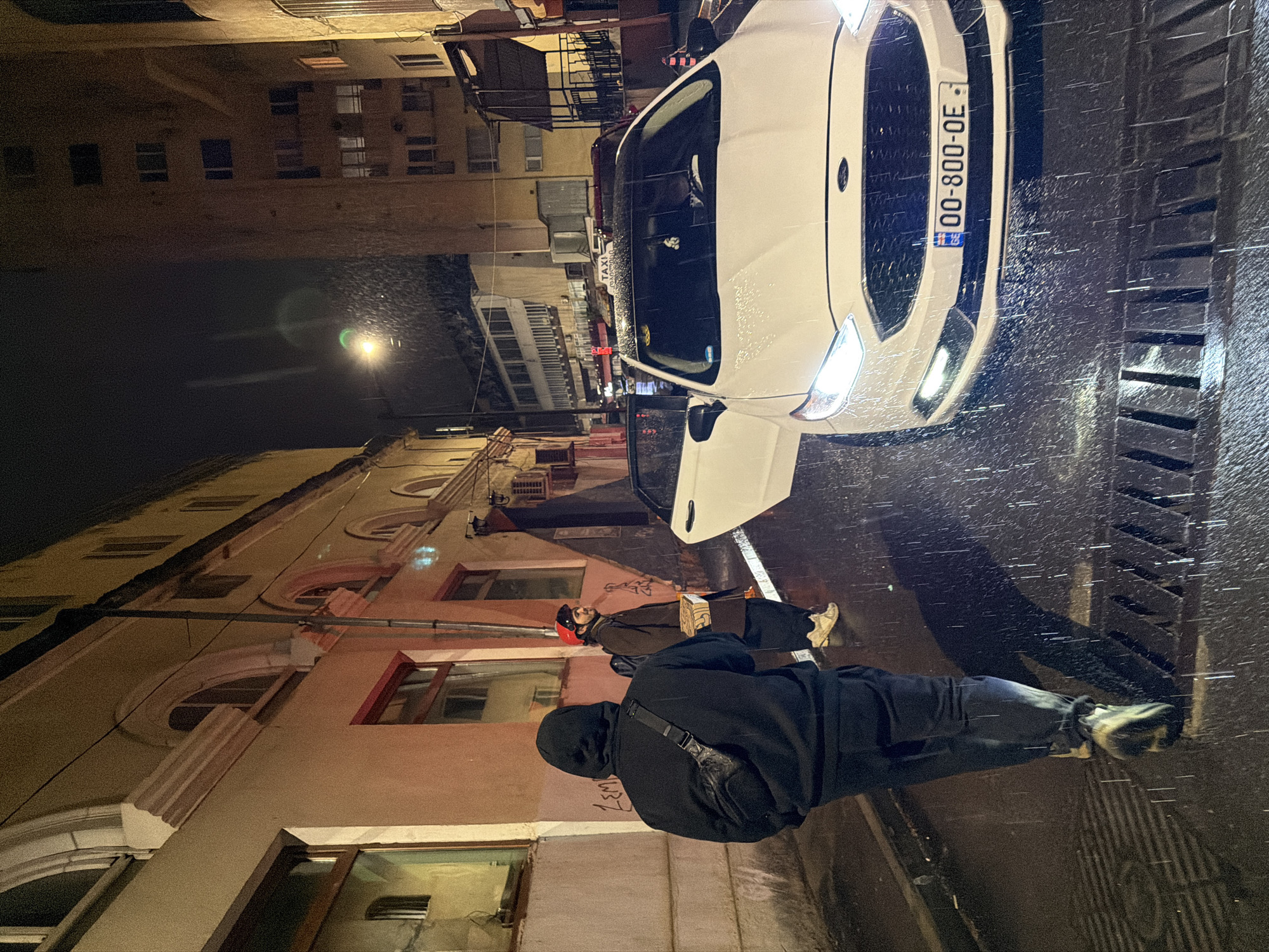 The image depicts a street scene at night, likely in Tbilisi, Georgia, as indicated by the Georgian license plate "30-800-OE" on the white Ford taxi. It appears to be raining. Two pedestrians are visible on the sidewalk: one wearing a dark hooded sweatshirt and carrying a shoulder bag and the other in a brown coat and red helmet. The taxi's driver-side door is open. The background shows buildings and street lighting. The wet street reflects the lights.