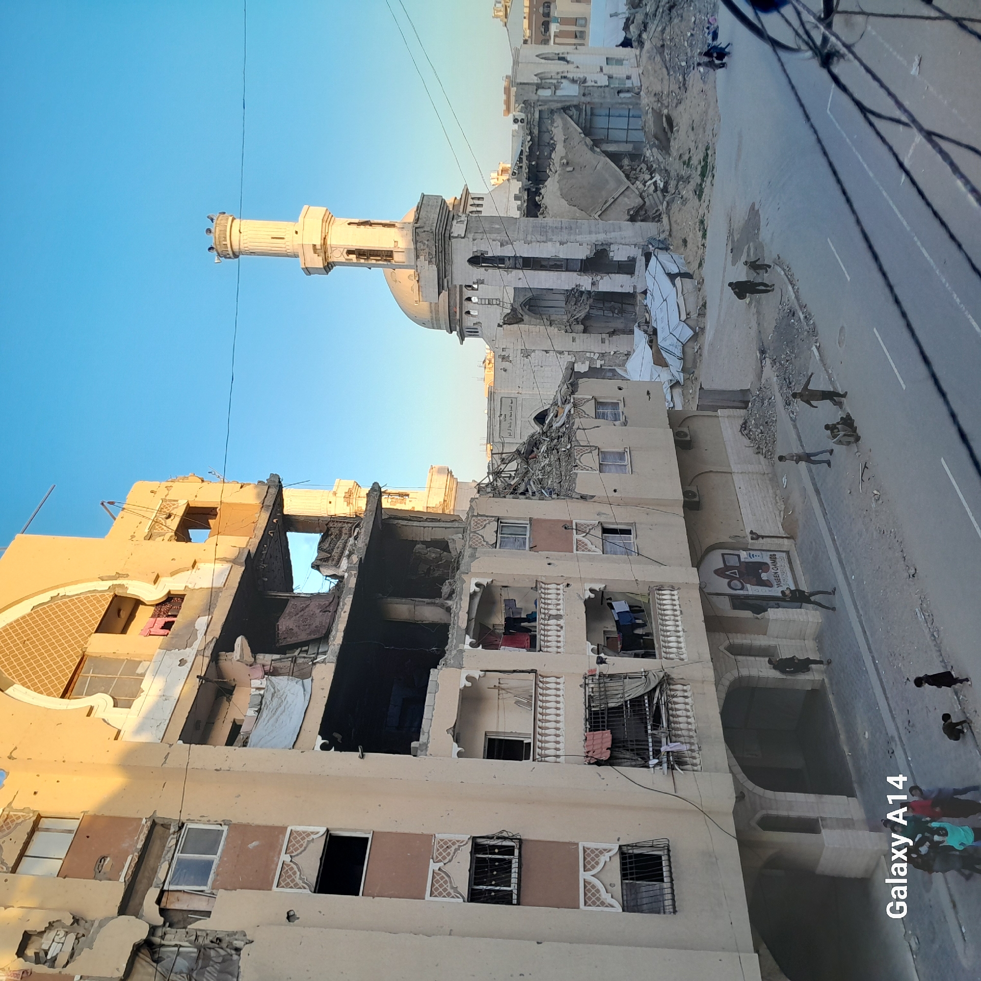 The image shows a cityscape in Unknown, Unknown, with a combination of destruction and daily life. In the foreground, a partially destroyed building reveals damaged interiors, including a room with bedding and remnants of household items. Clothes hang from windows. People are visible on the street, some walking and others grouped together. Further down the road, there's a partially ruined structure with a minaret. The background features additional buildings and a clear sky. A road cuts through the scene, separating the damaged buildings from the street activity. Overhead electrical wires are visible.