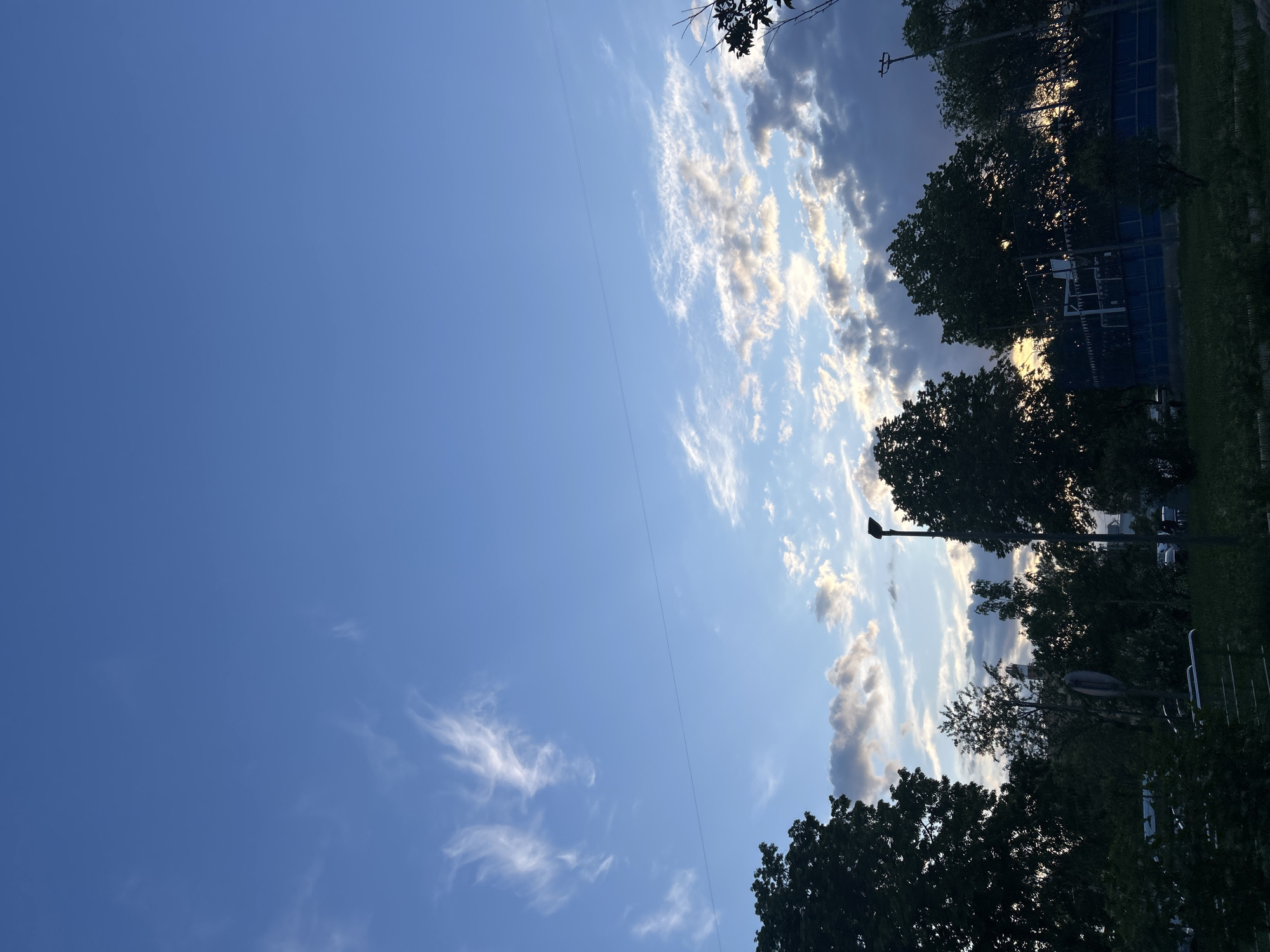The image, oriented vertically, primarily features a clear blue sky with various cloud formations. When viewed horizontally, the sky dominates the upper and left portions of the frame. In the upper left area, the sky is a deep, clear blue with sparse, wispy white clouds. Towards the center and right, more defined cumulus clouds appear, with some brightly illuminated by direct sunlight and others appearing darker grey due to shadow. A thin, dark wire, likely a power line, stretches diagonally across the sky.
Along the right and bottom-right edges of the image, a dense line of trees forms a dark silhouette against the brighter sky. Their foliage appears lush and full. The low angle of the sun, casting shadows and illuminating clouds from behind the trees, suggests either early morning or late afternoon. A tall, slender street light or utility pole is visible rising above the tree line. In the deeper background, behind the trees, dimly lit structures, possibly buildings or fences (some appearing with a blue hue), and patches of dark green grass are faintly discernible.
There are no people or specific activities taking place. No visible text is present in the image. The scene conveys an outdoor setting, likely a semi-urban or suburban area, characterized by natural elements and minimal infrastructure.