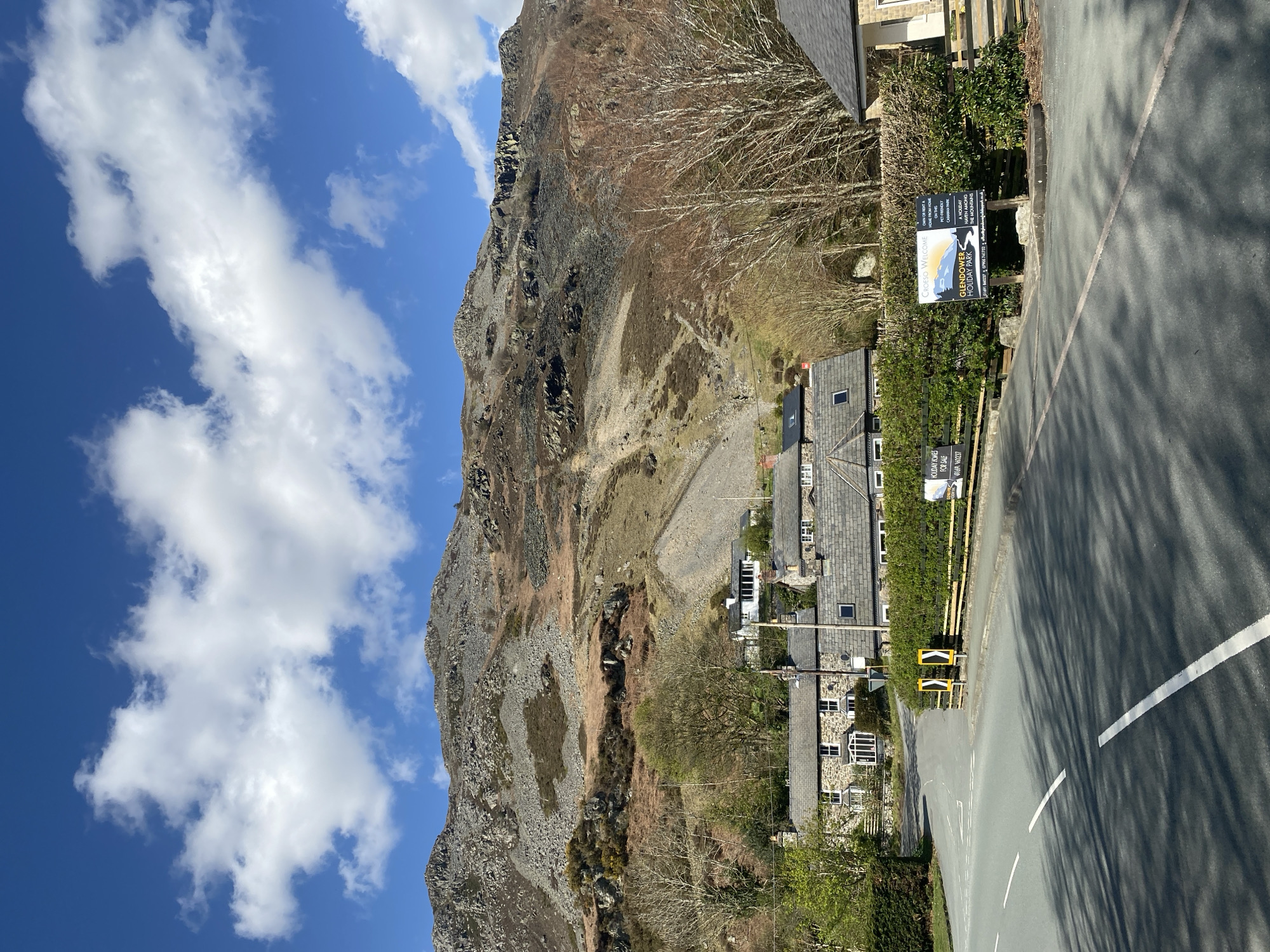 This image captures a view in Llangynog, United Kingdom. A towering slate mountain looms to the left, its rocky, quarried face visible against a backdrop of blue sky with scattered white clouds. A cluster of stone houses with slate roofs lines the roadside. A road runs along the side of the image with white lines and speed ramps are seen along the curb. A sign for the Glendower Holiday Park is affixed to a hedge along the road. The lower-left corner includes foliage, and long shadows are cast across the road.