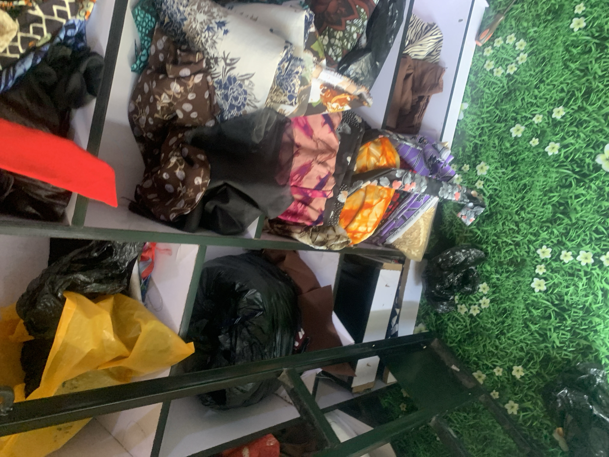A cluttered storage area in Jauro Yinu, Nigeria, contains fabric scraps and plastic bags. The shelves are filled with folded fabric of various patterns and colors, including floral, abstract, and animal prints, as well as solid colors. Several black plastic bags, some appearing to contain more items, are also present on the shelves and on the floor. A bright red piece of fabric is draped over one of the shelves. The floor is covered in a green mat designed to look like grass with small white flowers. A dark metal shelving unit is visible on the left side of the image.