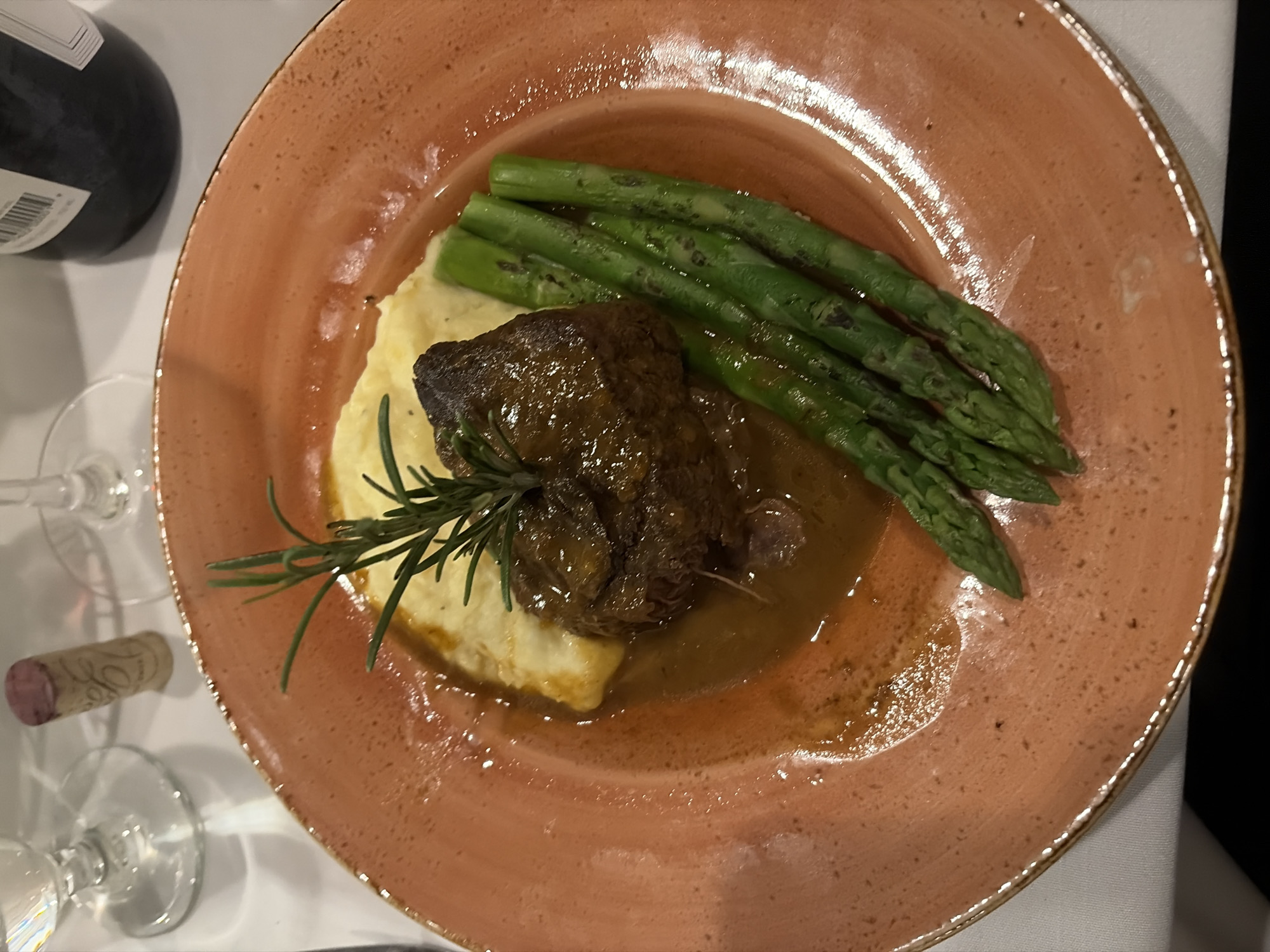 A close-up, overhead shot depicts a culinary dish presented on an orange, speckled plate. The meal consists of mashed potatoes topped with a braised meat item and garnished with a sprig of rosemary. A cluster of green asparagus spears is arranged adjacent to the meat and potatoes. A brown sauce or gravy partially surrounds the dish. In the background, a wine bottle, a cork, and stemware suggest a fine dining experience, but there is no evidence of a specific location within Key Biscayne, United States of America. There are no visible people.