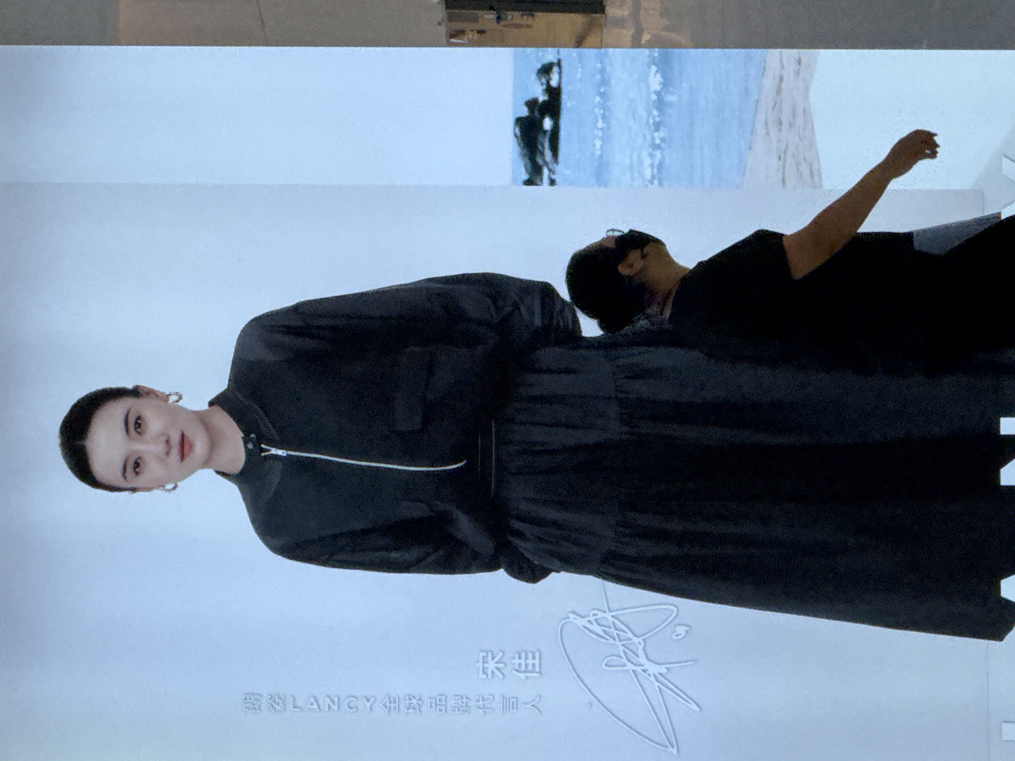 The image displays a poster featuring a woman in a black outfit, standing against a pale blue background. The woman has her hair pulled back, wears hoop earrings, and a faint smile. Text is visible on the left side of the poster, in both Chinese and English. Below the text is a white, stylized signature. To the right of the main subject, a second person, wearing a black shirt and a mask, is partially visible, looking to the right. The background transitions to a depiction of a beach with rocks in the water, suggesting an outdoor or scenic element. The overall impression is that of an advertisement or promotional material. The location is identified as Linghe, China.