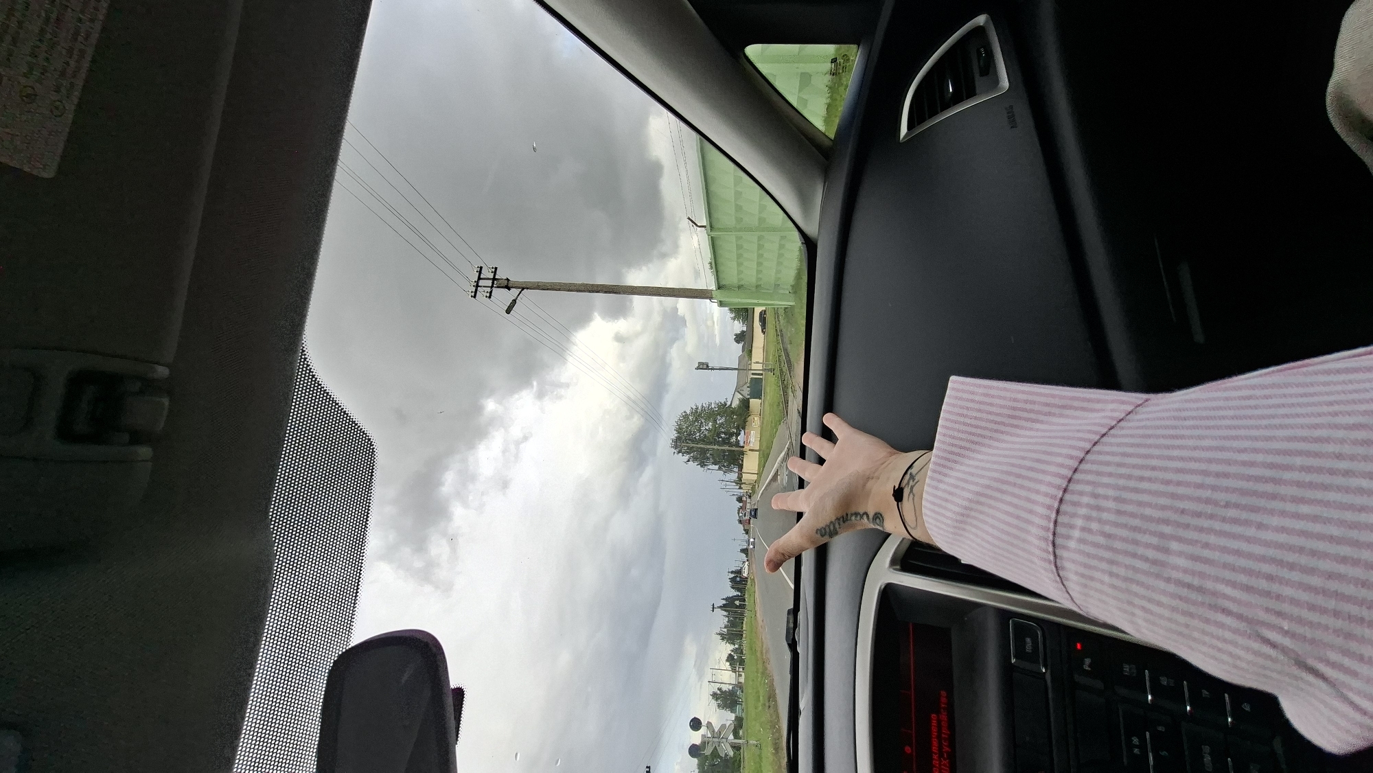 The image is taken from inside a car, looking out through the windshield. A person's arm and hand are visible in the foreground, extended towards the camera, with tattoos on the wrist. The person is wearing a pink and white striped shirt. The car is on a road with a cloudy sky overhead. Power lines and a utility pole are visible in the distance, as is a green fence. The road appears to be a rural or suburban area with some buildings and trees. The location is identified as Smalyavichy, Belarus.
