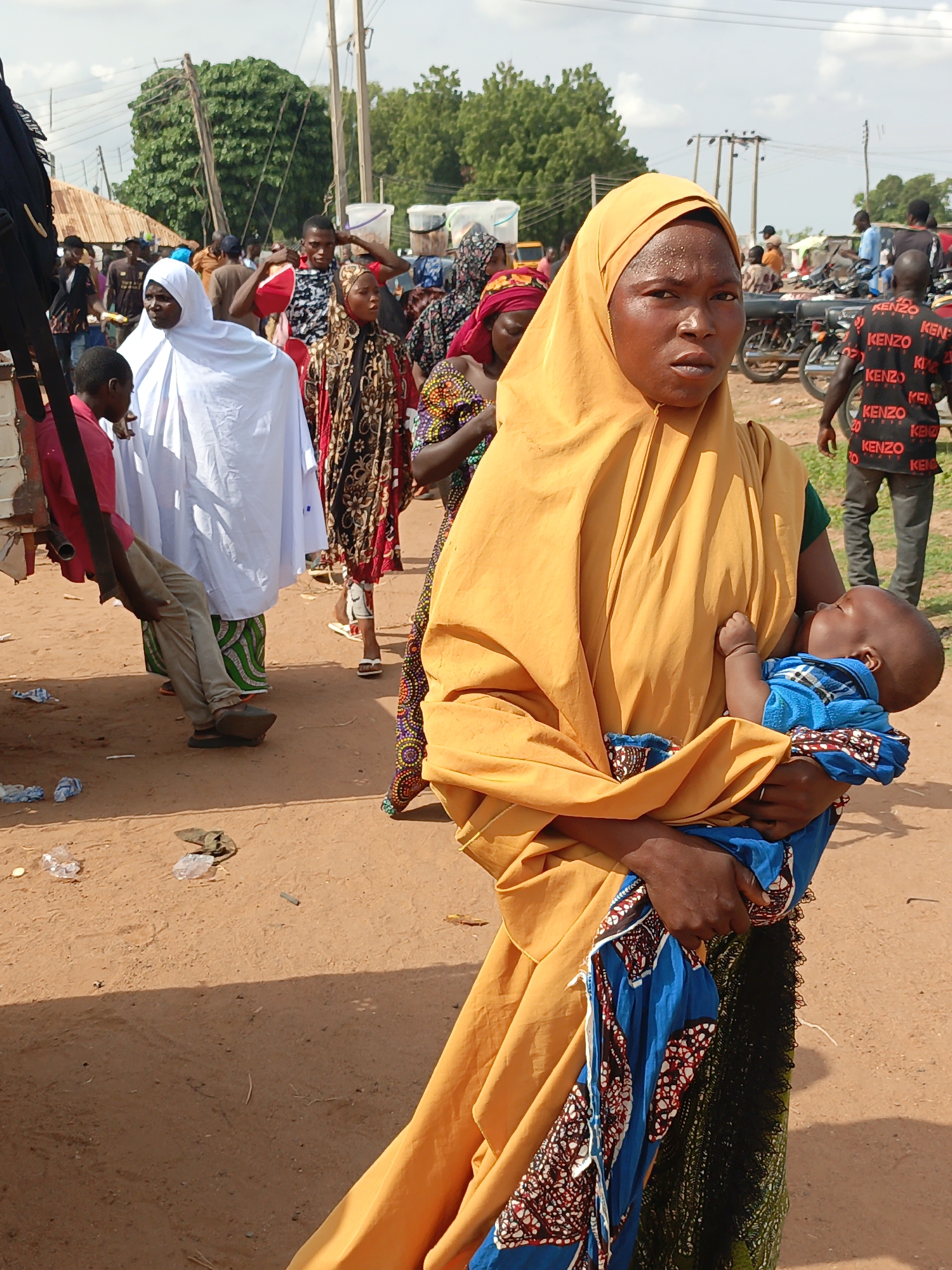 In a crowded area of Lafia, Nigeria, a woman with dark skin and an orange headscarf holds a baby wrapped in a blue patterned cloth. The woman looks directly at the camera. Other people are visible in the background, including women in headscarves and patterned clothing. There are motorcycles and buildings. The ground is dirt. Litter is scattered on the ground.