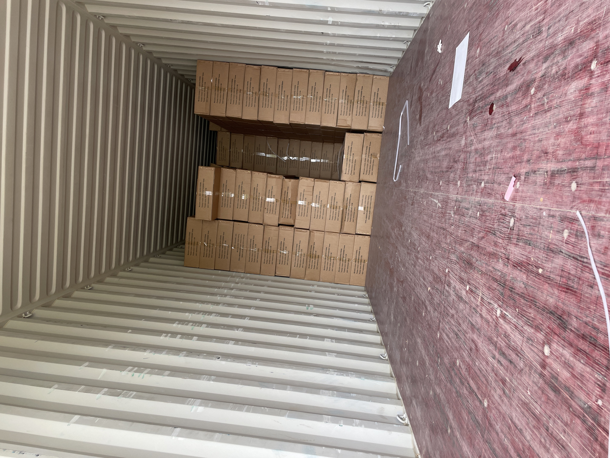 The interior of a standard shipping container is shown, loaded with stacks of cardboard boxes. The container walls are corrugated metal. The container's floor is also metal. The boxes appear to be similarly sized and arranged in neat rows, reaching approximately halfway up the container's height. The wooden rear wall of the container is visible, with scattered markings and residue. The image does not contain any information regarding the city of Asenovgrad, Bulgaria. There are no people visible in the image.