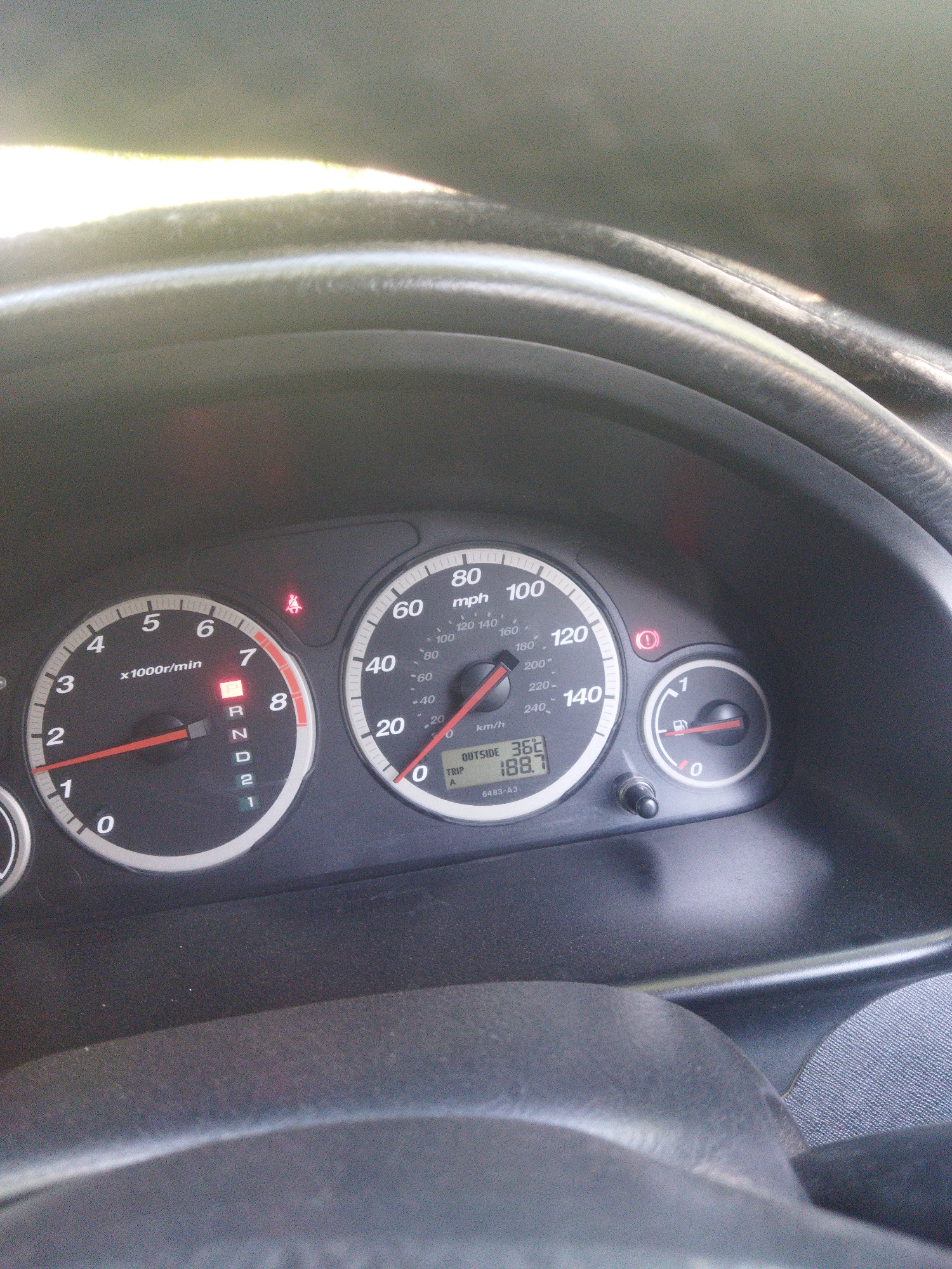 The image shows a close-up of a car's dashboard. A speedometer displays a speed of 0 mph, and a tachometer shows an RPM of approximately 700. The gear selector is in the "R" (Reverse) position. To the right of the speedometer, there is a fuel gauge showing nearly empty, and an outside temperature display indicating 36 degrees. A small digital display shows "TRIP A 188.7". There is also a seatbelt warning light illuminated. The dashboard is dark gray and appears to be in a car in Mailuu-Suu City, Kyrgyzstan.