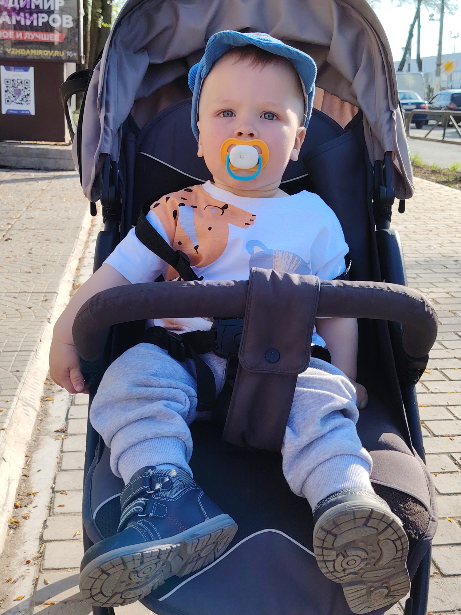 A toddler wearing a blue hat, a white T-shirt with an orange animal print, and gray sweatpants is seated in a dark gray stroller with a brown handlebar and a black safety harness. The child has a pacifier in their mouth and is wearing navy blue shoes. The stroller is on a brick sidewalk. Behind the stroller, there is a building with a sign in Russian "ВЛАДИМИР АМИРОВ ВОЕ И ЛУЧШЕЕ VZHDAMIROV.RU 16+", as well as a QR code and telephone number. Further in the background, there is a street with parked cars and trees. The image was taken in the city of Chaikovsky, Russia.