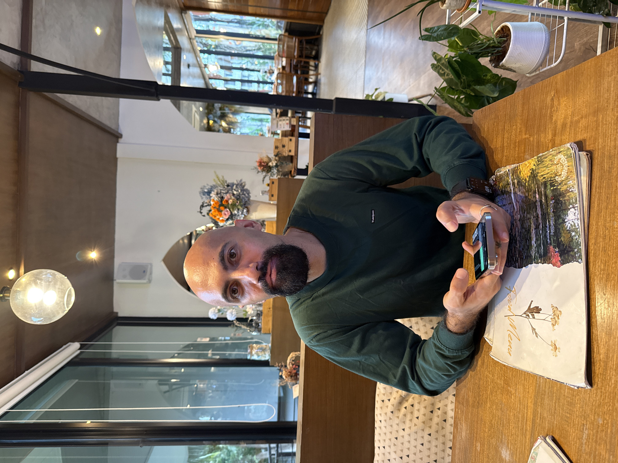 A man with a bald head and beard sits at a wooden table in what appears to be a restaurant or cafe. He is wearing a dark green long-sleeved shirt. In front of him is a menu with a floral design. He is holding a smartphone in his hands, looking down at it. The background shows a dining area with large windows revealing greenery outside, tables, and chairs, suggesting a location in Ko Samui District, Thailand. Decorative elements such as vases of flowers and plants are visible.