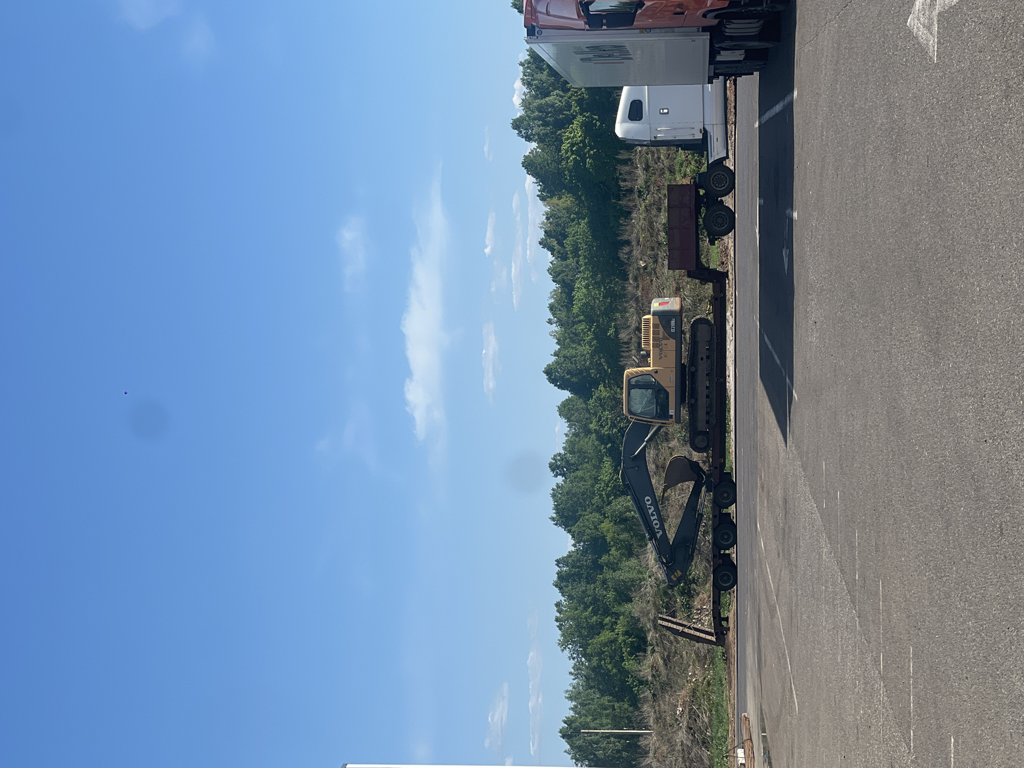 A parked excavator on a trailer behind a semi truck in Kazan, Russia, adjacent to a wooded area and a paved parking lot under a blue sky with scattered clouds.