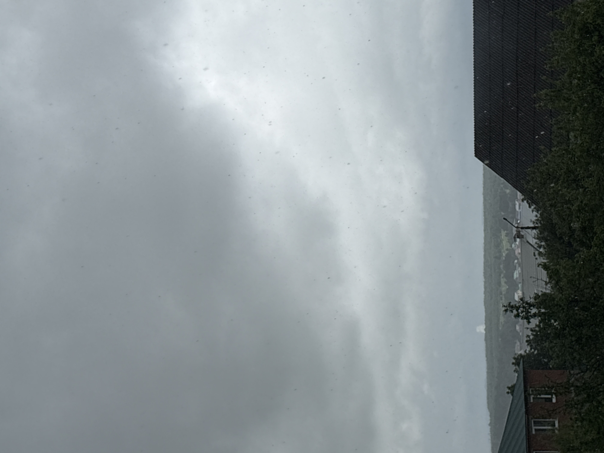 The image shows a cloudy, overcast sky above a scene in Прикамский, Russia. In the lower right corner, there is a section of a brick building with windows, partially obscured by a green leafy tree. Adjacent to the brick building, a dark-colored building or structure is visible, with what appears to be a dark roof and vertical siding. Beyond these buildings is a glimpse of the city landscape, possibly a row of buildings or structures along a distant line. The primary focus is the heavy cloud cover, suggesting a rainy or snowy condition. Small, light-colored dots, likely snowflakes, are visible in the air.