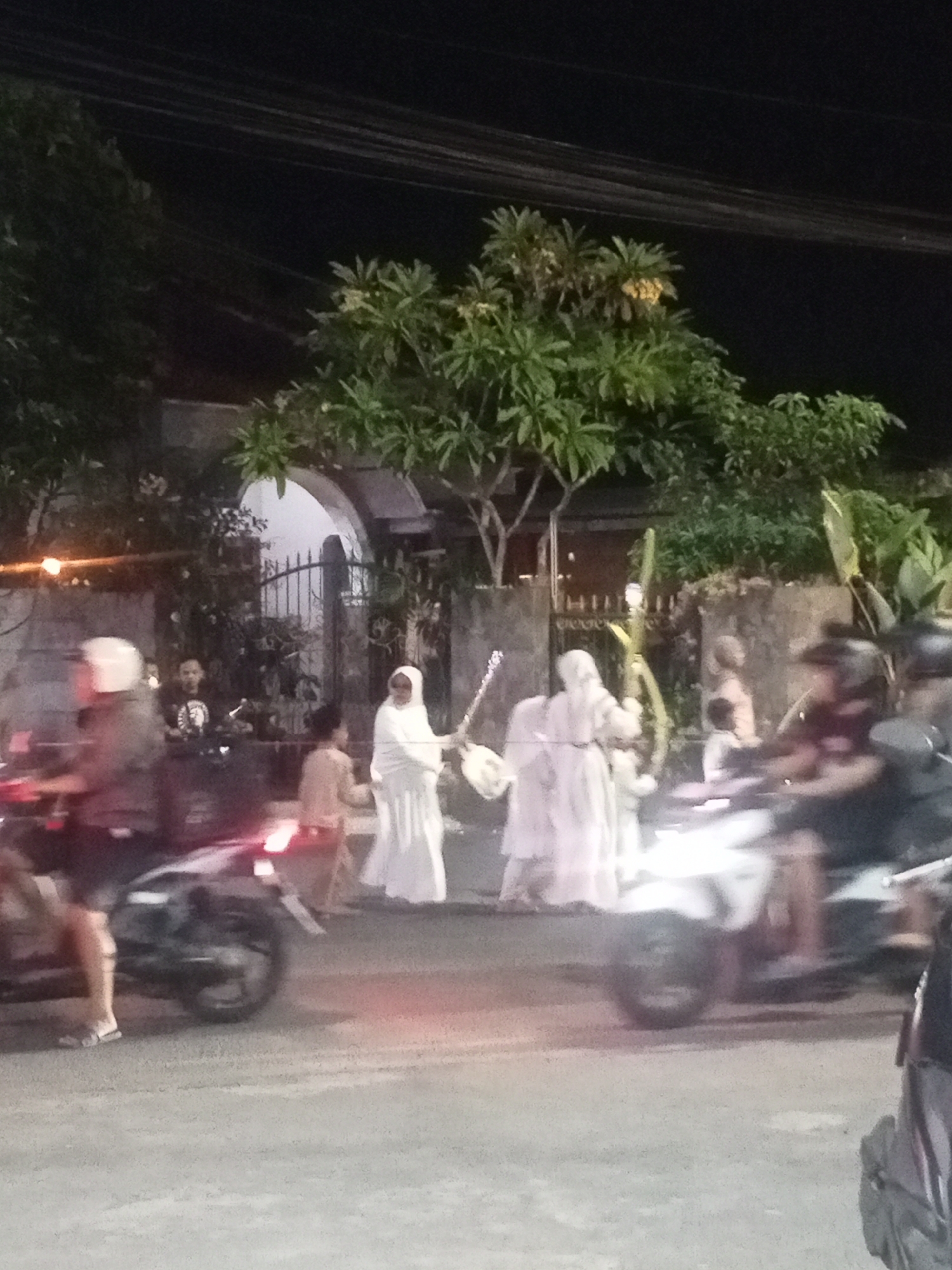 In Jember, Indonesia, at night, a group of people are seen near an arched gateway with a decorative fence. Several individuals are dressed in white garments, with some appearing to be wearing hijabs. One person in white is holding what looks like a musical instrument, possibly a stringed instrument, and another person is holding a long, curved object. Some people are on motorcycles, with one individual in the foreground wearing a white helmet. The scene takes place on a paved road, with trees and foliage visible in the background. The lighting suggests a streetlamp or ambient light from nearby buildings illuminating the area.