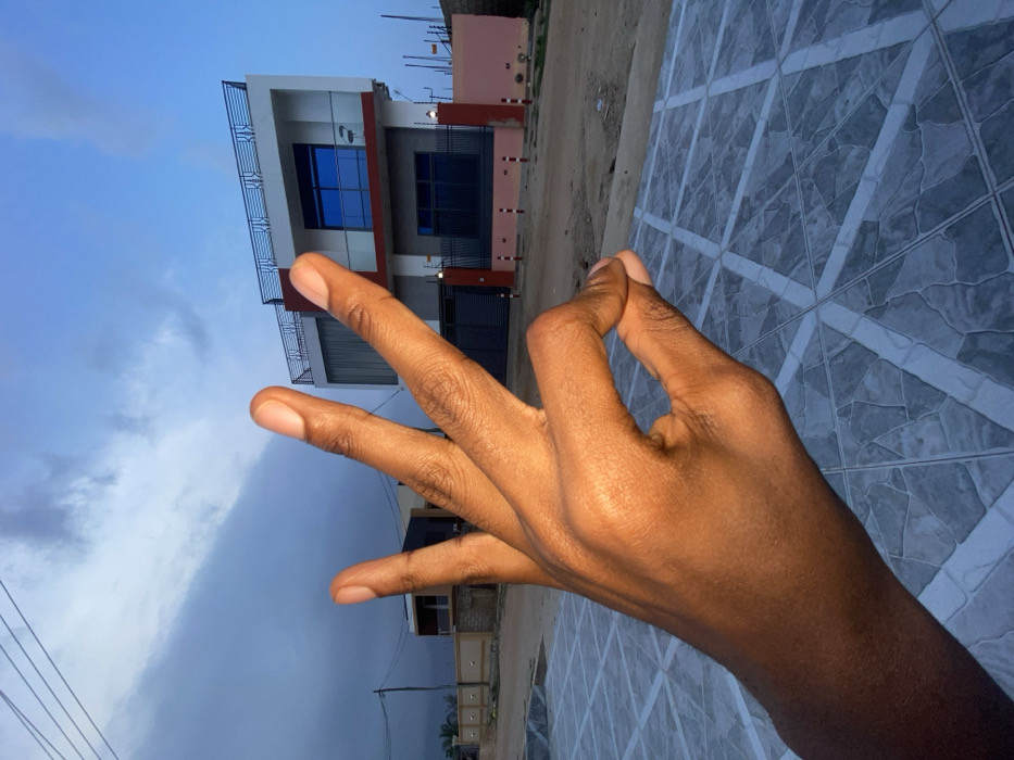 A hand, palm towards the camera, displays a hand gesture in front of a building and sky. Three fingers extend upward, while the thumb and pinky are curled together. The building, two stories, features a white facade, blue windows, and a red accent stripe. A cloudy sky is visible behind the building. A tiled surface with a geometric pattern fills the right foreground.