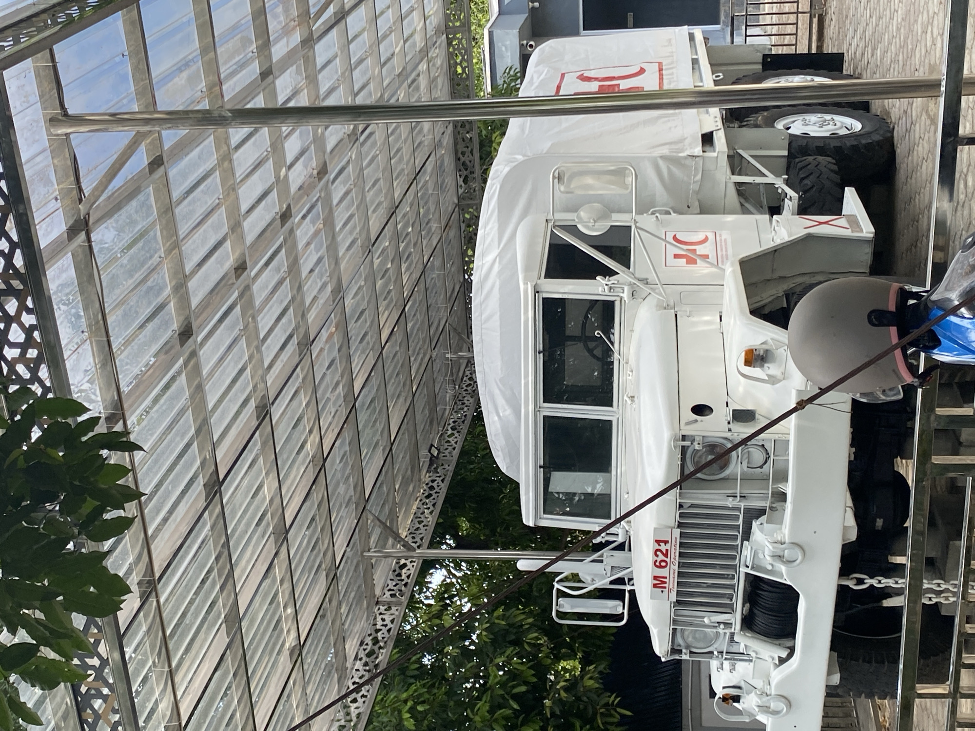 A white, vintage military truck, identified by the license plate "M 621 Tsunami Operation," is parked under a transparent corrugated roof structure. The truck features a winch on its front bumper and has "International Red Cross" symbols on its side. The vehicle appears to be in good condition, suggesting it is a preserved artifact or monument. The scene is set in an outdoor environment, likely a museum or memorial site in Banda Aceh, Indonesia, as indicated by the historical context of the "Tsunami Operation" designation. The background shows lush green foliage and part of a building. The lighting suggests it is daytime, with a slightly overcast sky filtering through the roof. There are no people prominently visible in the image.