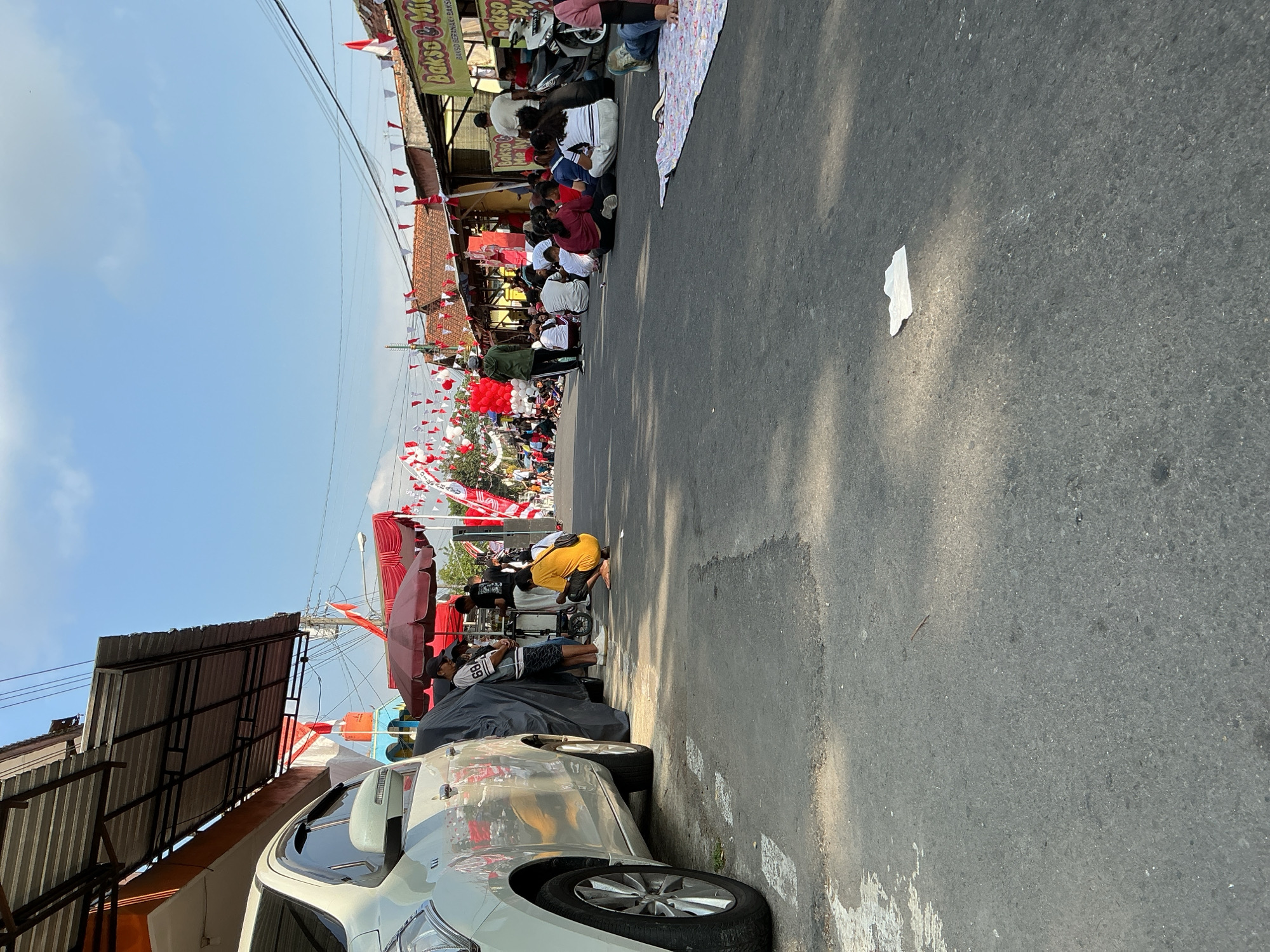 The image shows a street scene in Magelang, Indonesia, with a clear blue sky and scattered clouds. The street is decorated with numerous red and white Indonesian flags and pennants, suggesting a national celebration or event. Several people are present, some sitting on the ground, while others stand or move about. A white car is parked on the left side of the road, partially covered by a dark tarp. Several roadside stalls or shops are visible on the right, with signs indicating food vendors, one reading "Bakso & Mie" (Meatball & Noodles). A red umbrella provides shade for some individuals near the car. A person in a yellow shirt is crouching on the road, and another person in a green jacket is standing in the distance. Several individuals are sitting on blankets and directly on the asphalt, facing the direction of the stalls. The overall atmosphere appears to be a community gathering or market.