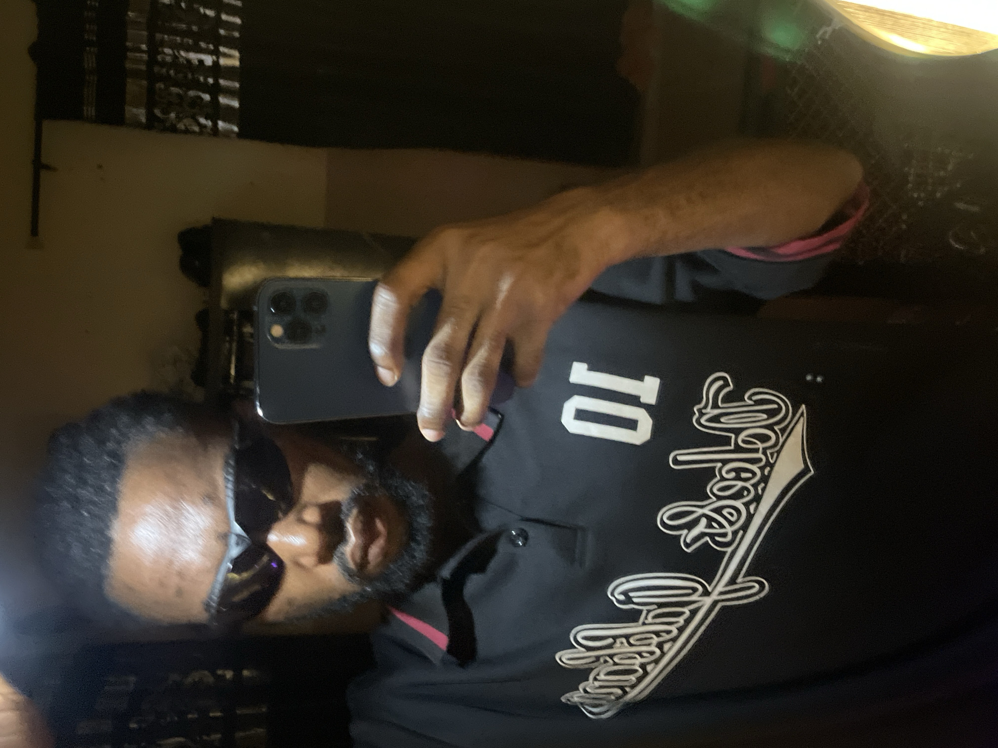 A Black male, appearing to be in his 20s or 30s, with short dark hair and a beard, is depicted taking a mirror selfie indoors in Lokoja, Nigeria. He is wearing dark wraparound sports-style sunglasses and a black polo shirt with a red trim on the collar and sleeve cuffs. On the right side of the chest (viewer's left in the reflection), a white embroidered number "10" is prominently displayed. On the left side of the chest (viewer's right in the reflection), white embroidered cursive text is visible, appearing horizontally reversed due to the mirror image. The subject holds a dark-colored smartphone, likely an iPhone with multiple rear cameras, in his right hand, which is visible from the wrist upwards. The background, visible in the mirror's reflection, shows a dimly lit room with light-colored walls and dark patterned curtains on the right side of the frame. A dark piece of furniture with objects on top is partially visible on the far left.